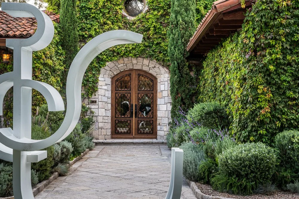 A garden pathway leading to a wooden door with ironwork, surrounded by lush green foliage and plants, with part of a modern sculpture in the foreground. San Miguel de Allende luxury real estate photography. Vinedos San Lucas