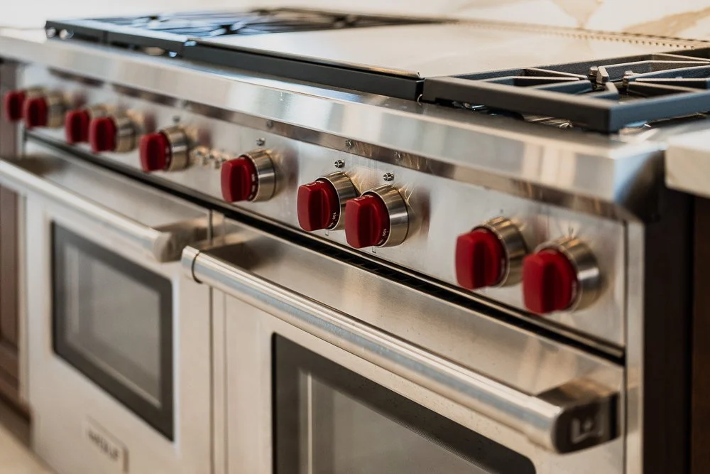 Stainless steel stove with six red knobs and two oven doors below in a kitchen.