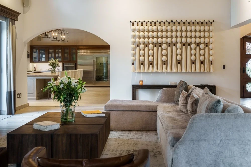 Living room with beige sectional sofa, wooden coffee table with books and a vase of flowers, wall art with wooden balls and tassels, and an open view into the kitchen and dining area. San Miguel de Allende luxury real estate photography.