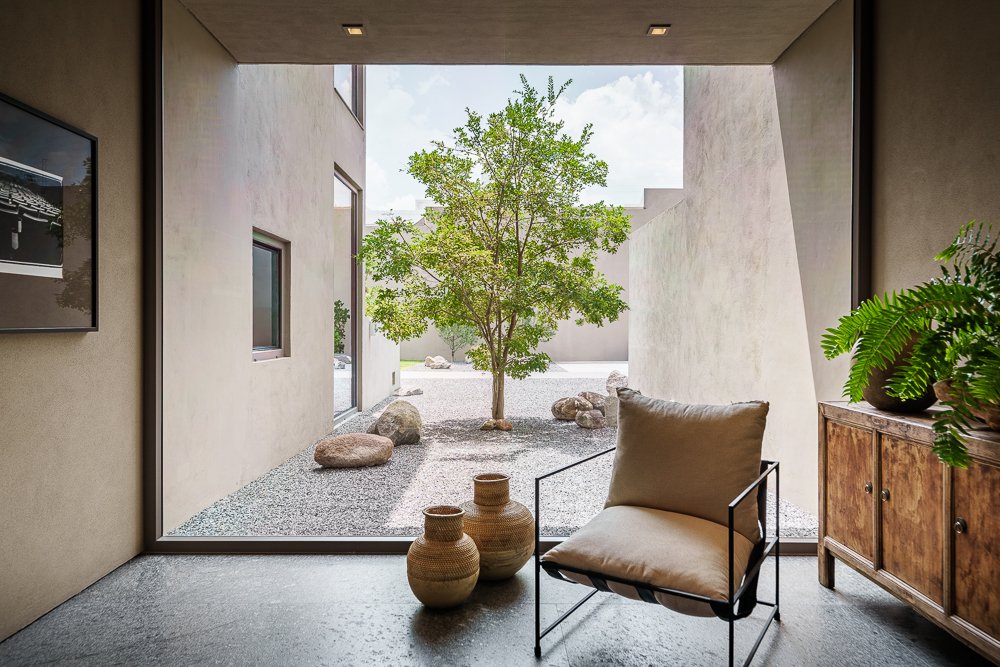 San Miguel de Allende luxury real estate photography. Interior view of a room with a large window looking out to a minimalist courtyard with a small tree and rocks, a beige cushioned chair, woven vases, and potted plants.