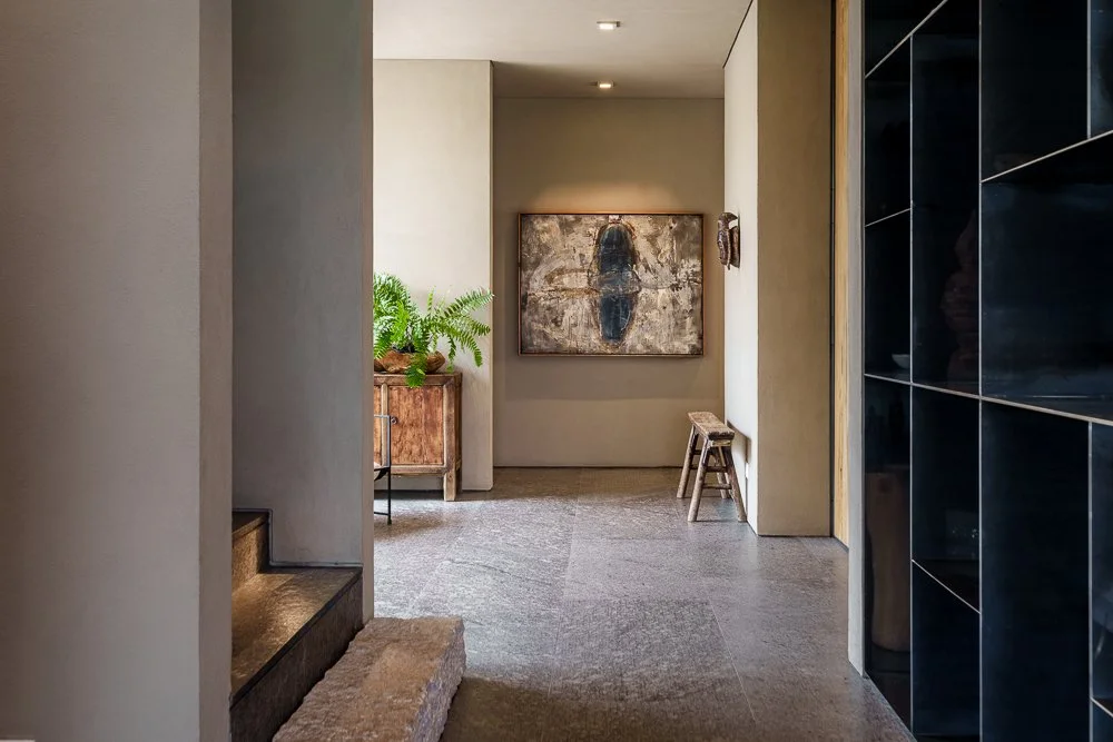 Interior view of a modern home hallway with artwork, a wooden bench, a cabinet with green plants, and black shelving. San Miguel de Allende luxury real estate photography.