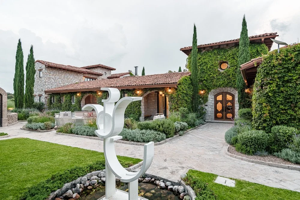 A luxurious house with stone walls and lush green plants, including tall cypress trees, and a modern sculpture in the front yard. San Miguel de Allende luxury real estate photography.