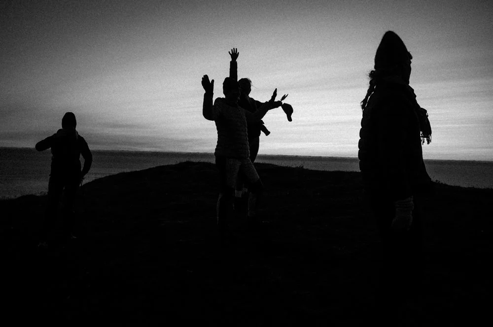 Silhouettes of five people on a beach at sunset, some with arms raised, wearing jackets and hats.