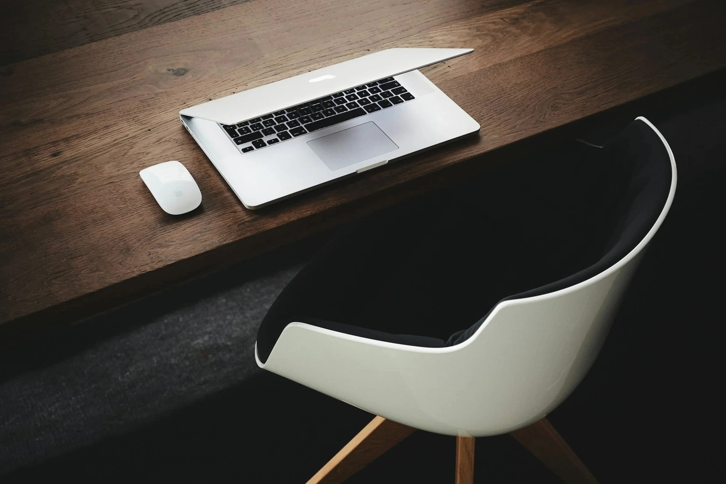 Wooden desk with an open MacBook, a wireless mouse, and a modern chair with a black seat and white shell design.