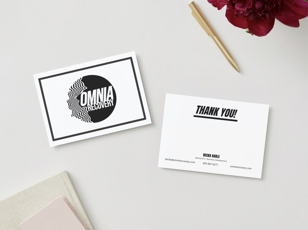 A thank you card and a business card on a white surface, with a gold pen, a pink notebook, and a pink flower in the background.