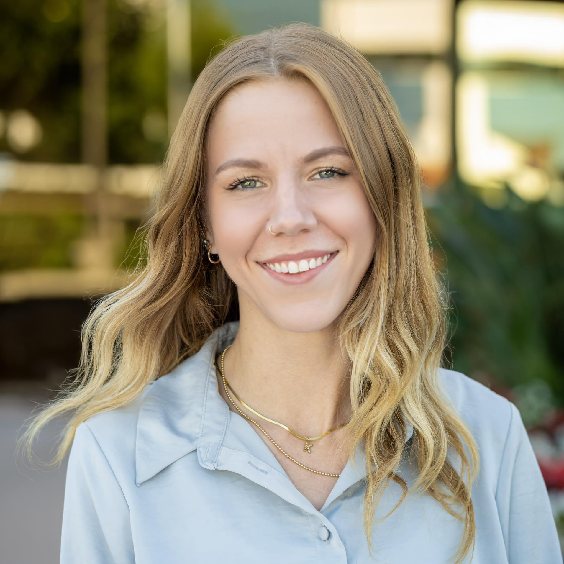 Smiling young woman with wavy blonde hair, wearing a light blue button-up shirt, gold jewelry, and a nose ring, outdoors with green foliage and sunlight in the background.