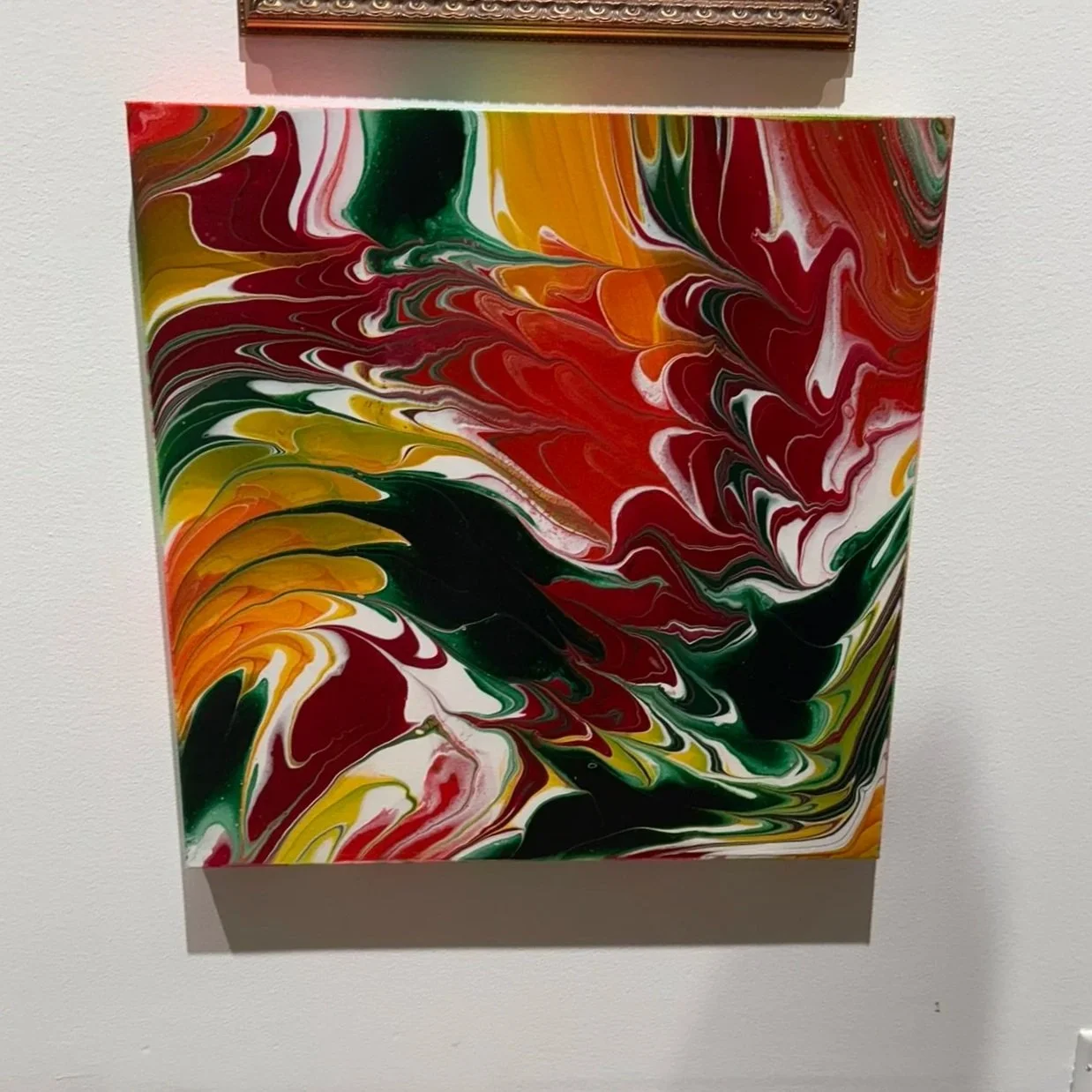 Abstract painting with swirling patterns of red, green, yellow, orange, black, and white colors on a canvas mounted on a white wall.