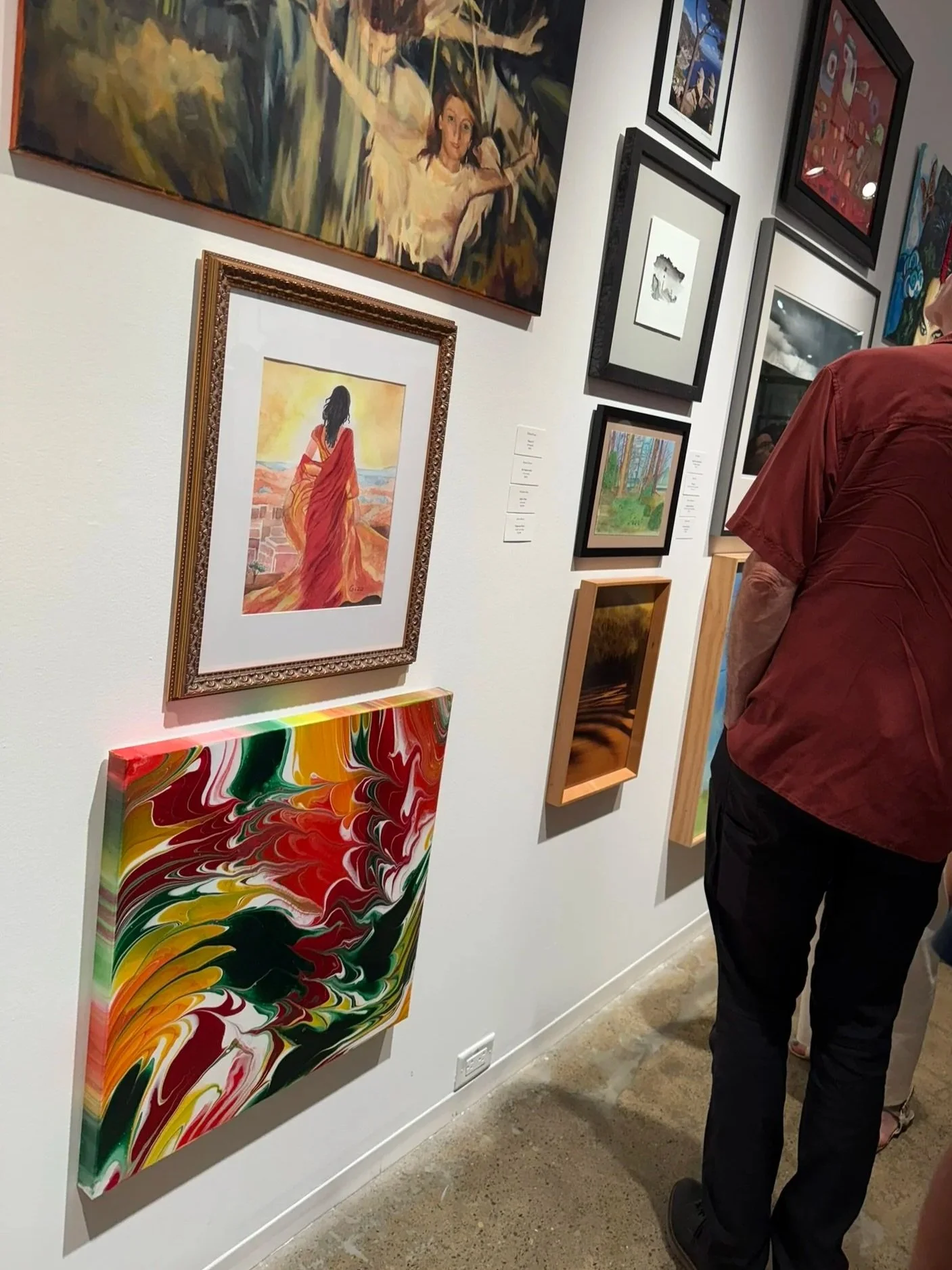 Gallery wall with various paintings including a woman in a red dress, abstract colorful art, a landscape, and other artwork, with a person in a red shirt viewing it.