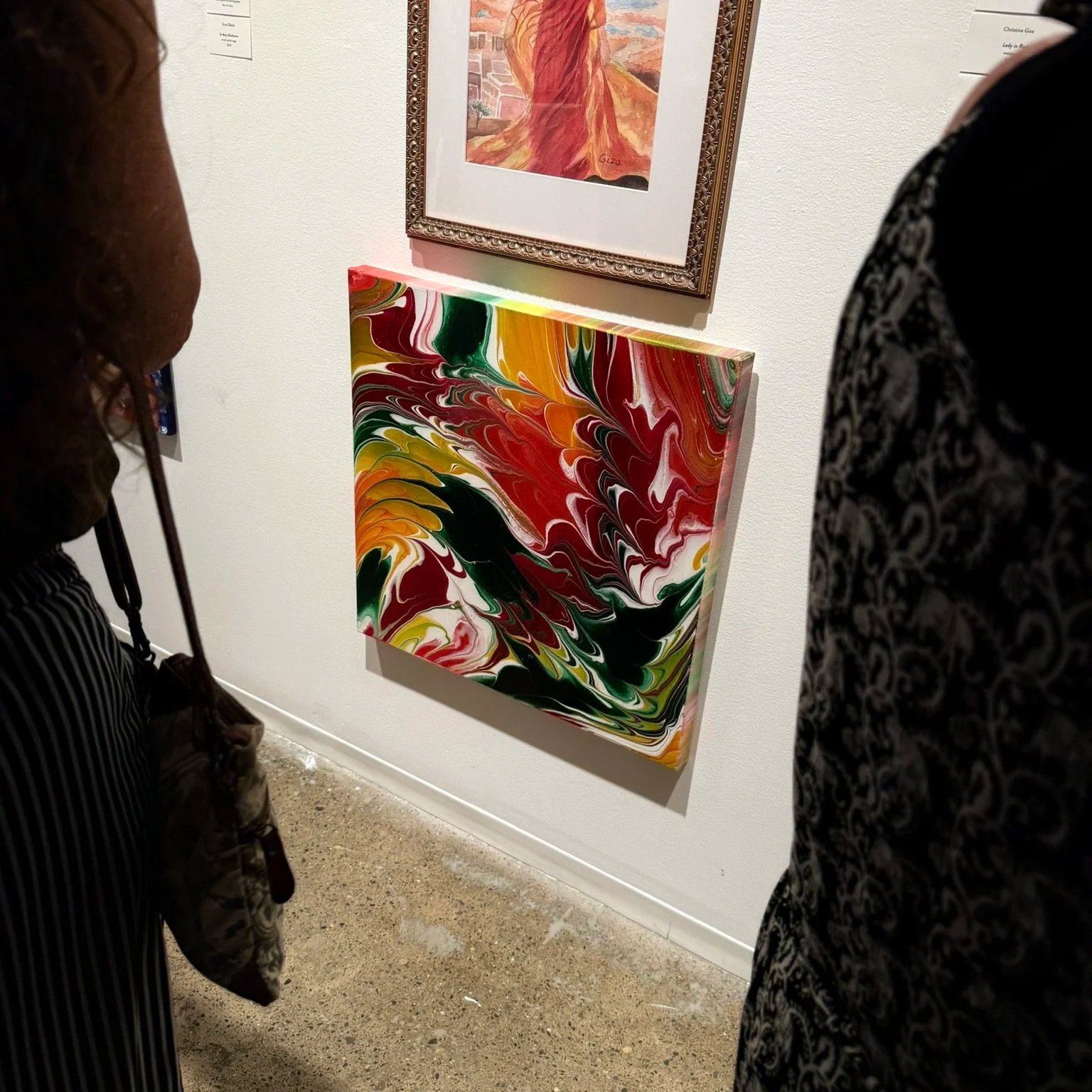 Two people are observing a colorful abstract painting in an art gallery. The painting features swirling patterns of red, yellow, green, black, and white.