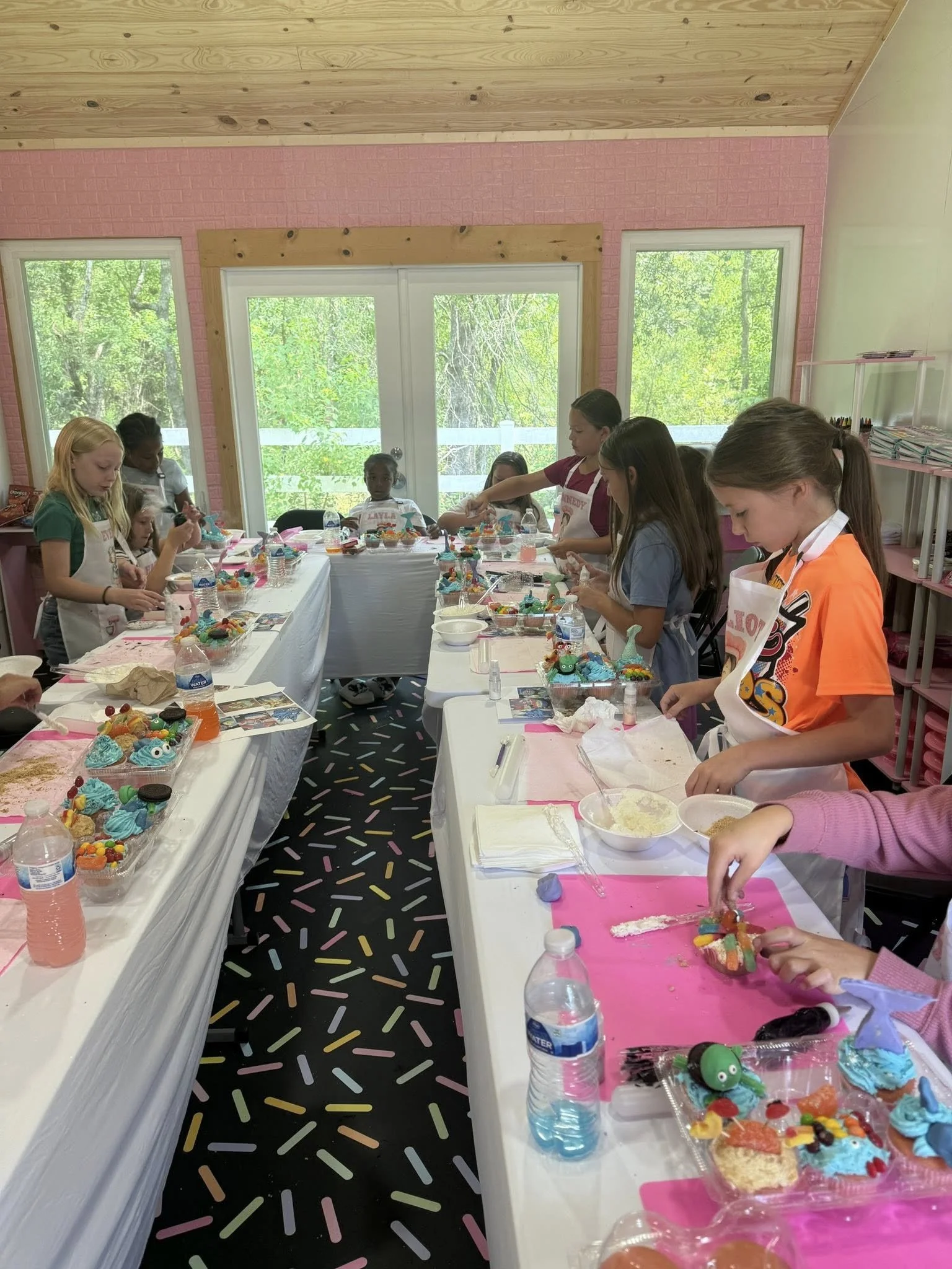 Children participating in a cake decorating activity at a baking class, with tables covered in pink and white, decorated cupcakes, and ingredients on the tables.