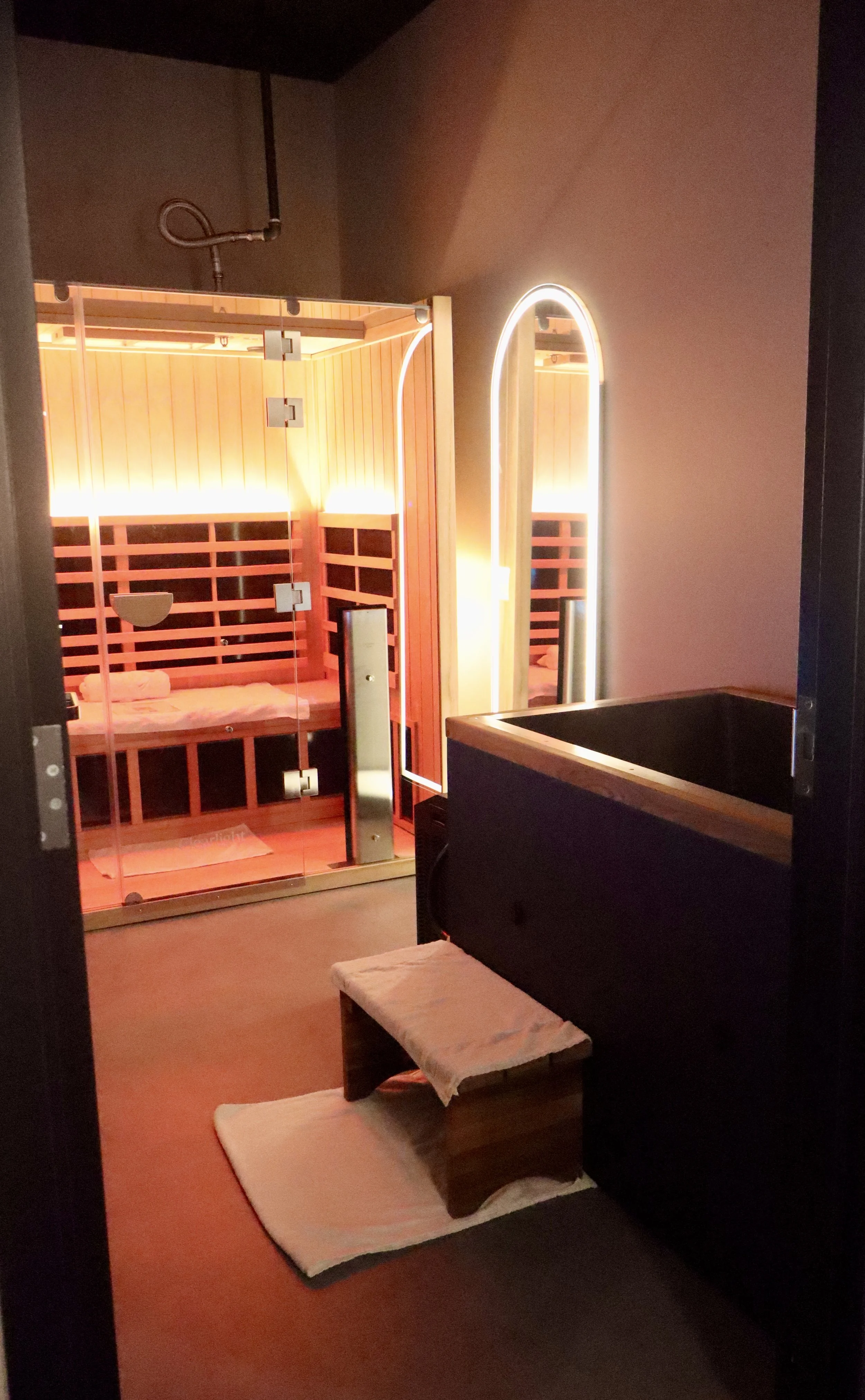 A sauna room with wooden interior, glass door, and a bench inside, with a heated space and modern lighting. Outside the sauna, there is a wooden hot tub with a towel-covered step.