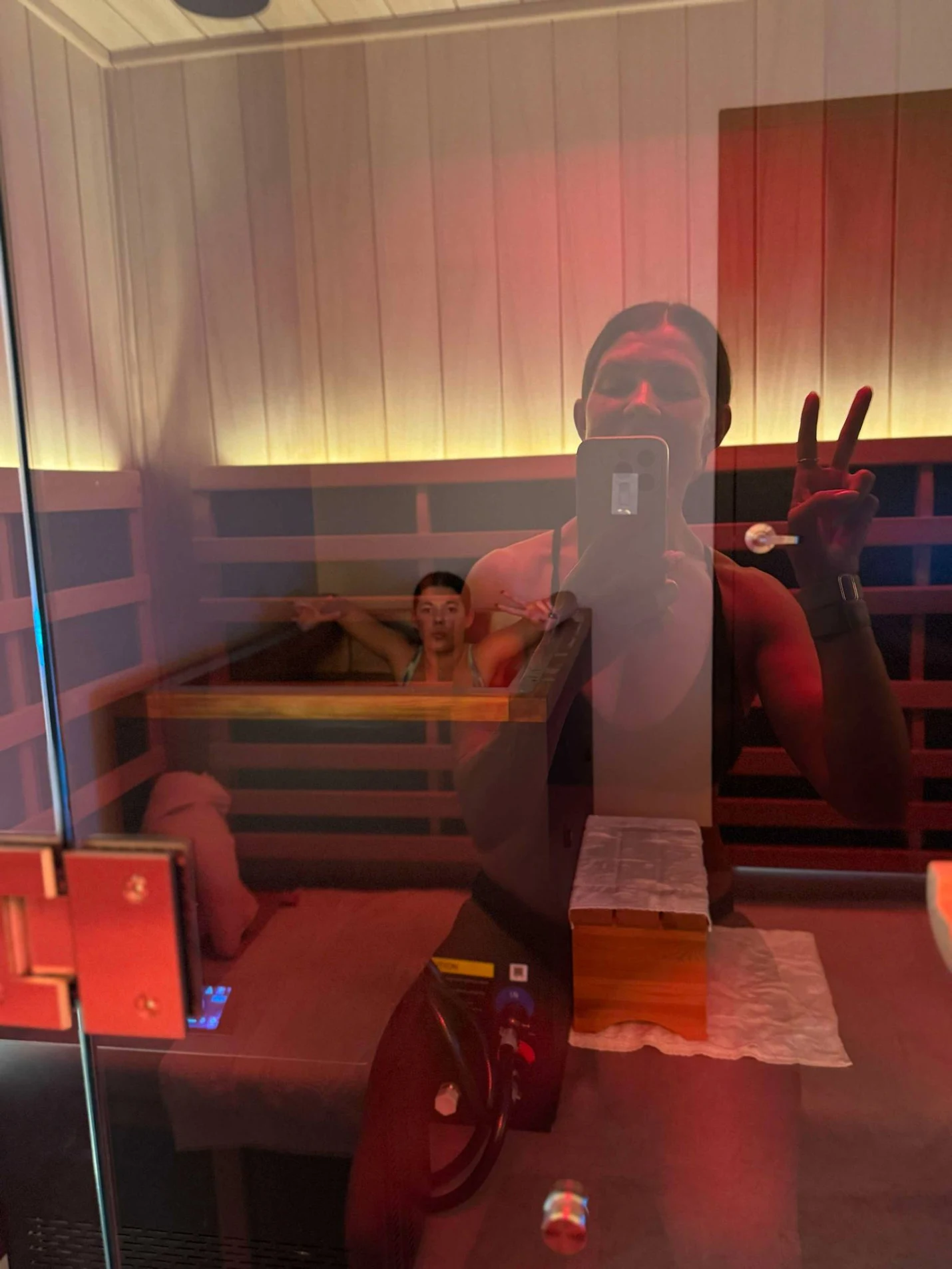 Reflection of two women in a glass door: one woman is inside a sauna with arms outstretched, and another woman is taking a selfie outside the sauna, making a peace sign with her hand.
