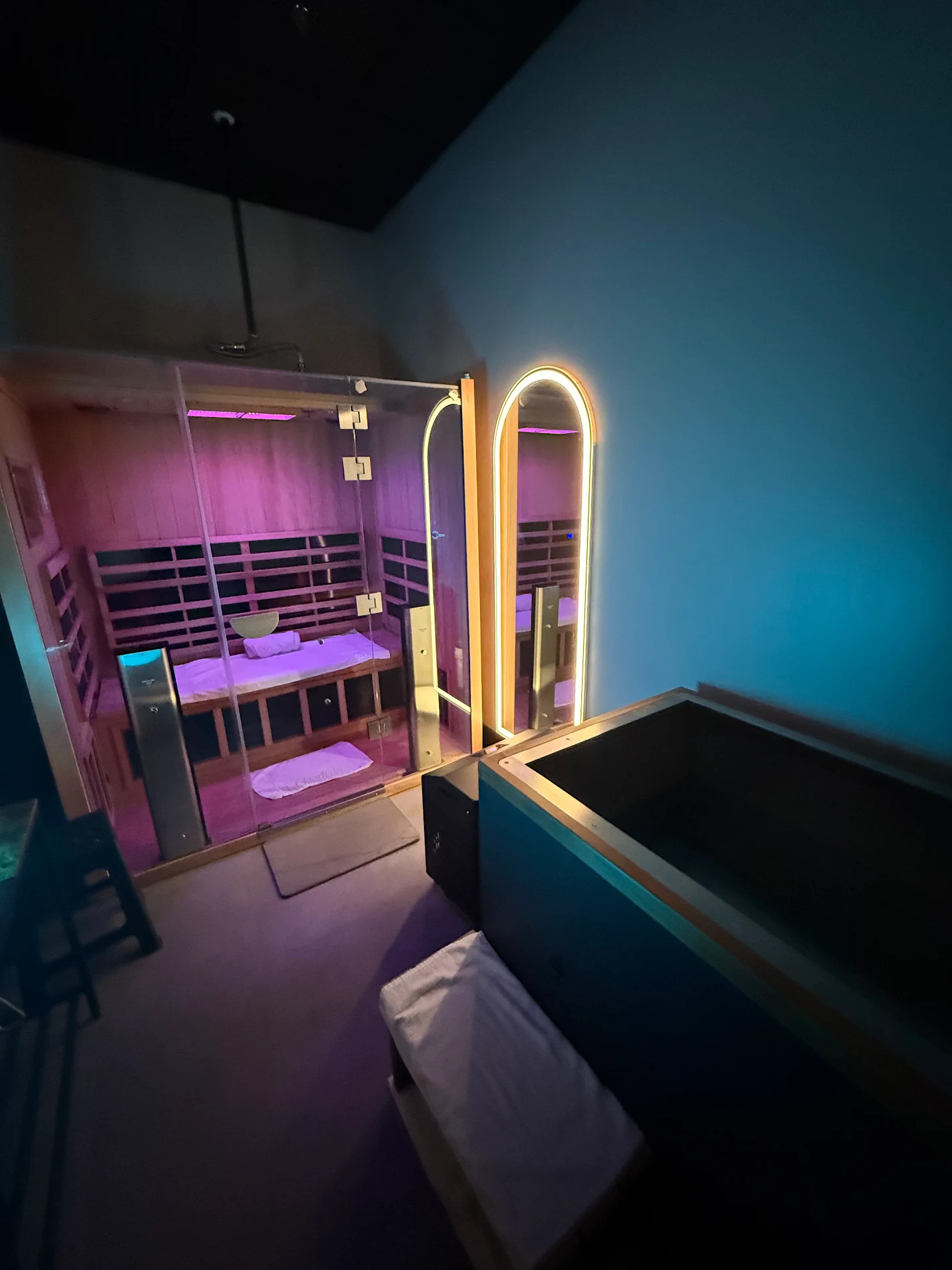 A room with a glass-enclosed sauna and a hot tub. The sauna is lit with pink and purple LED lights and has bunk beds inside. The hot tub is on the right side of the image, with a white towel placed on a seat nearby.