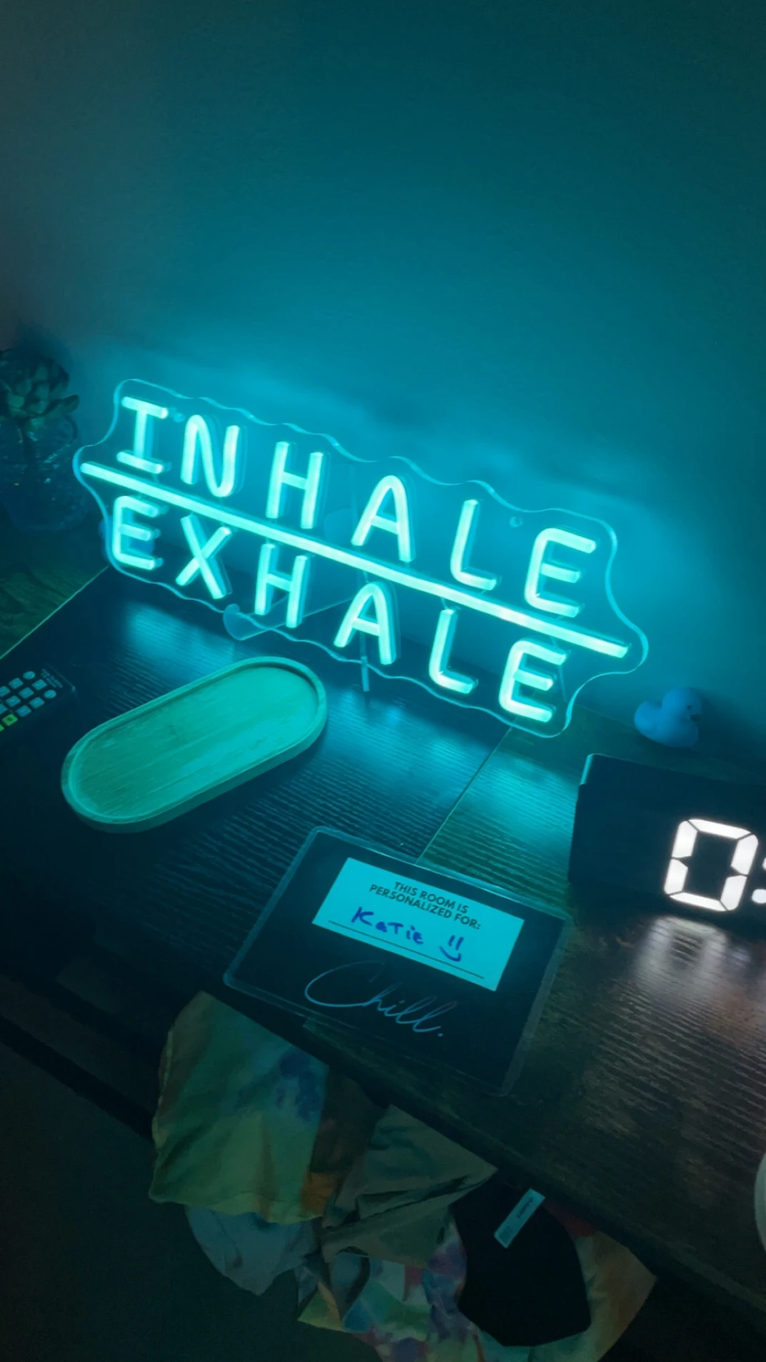 Neon sign with the phrase "Inhale, Exhale" on a table. There is a small plants, a remote, a tray, a tablet, and a digital clock on the table.
