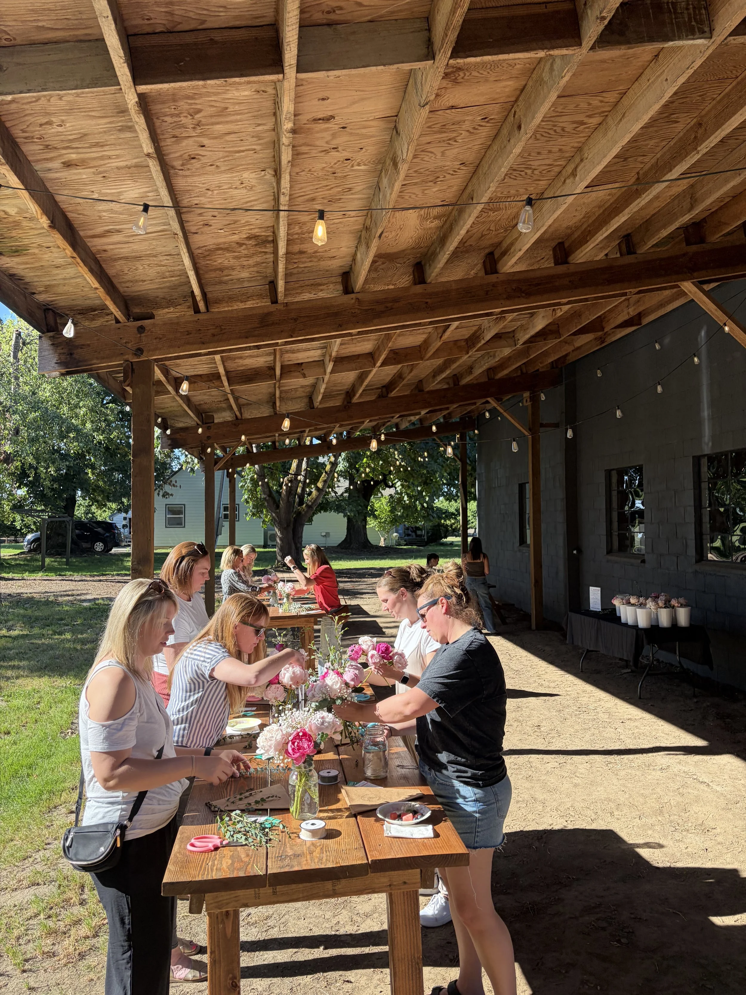 People arranging pink and white flowers on a wooden table outside under a wooden roof, with string lights hanging above and trees in the background.