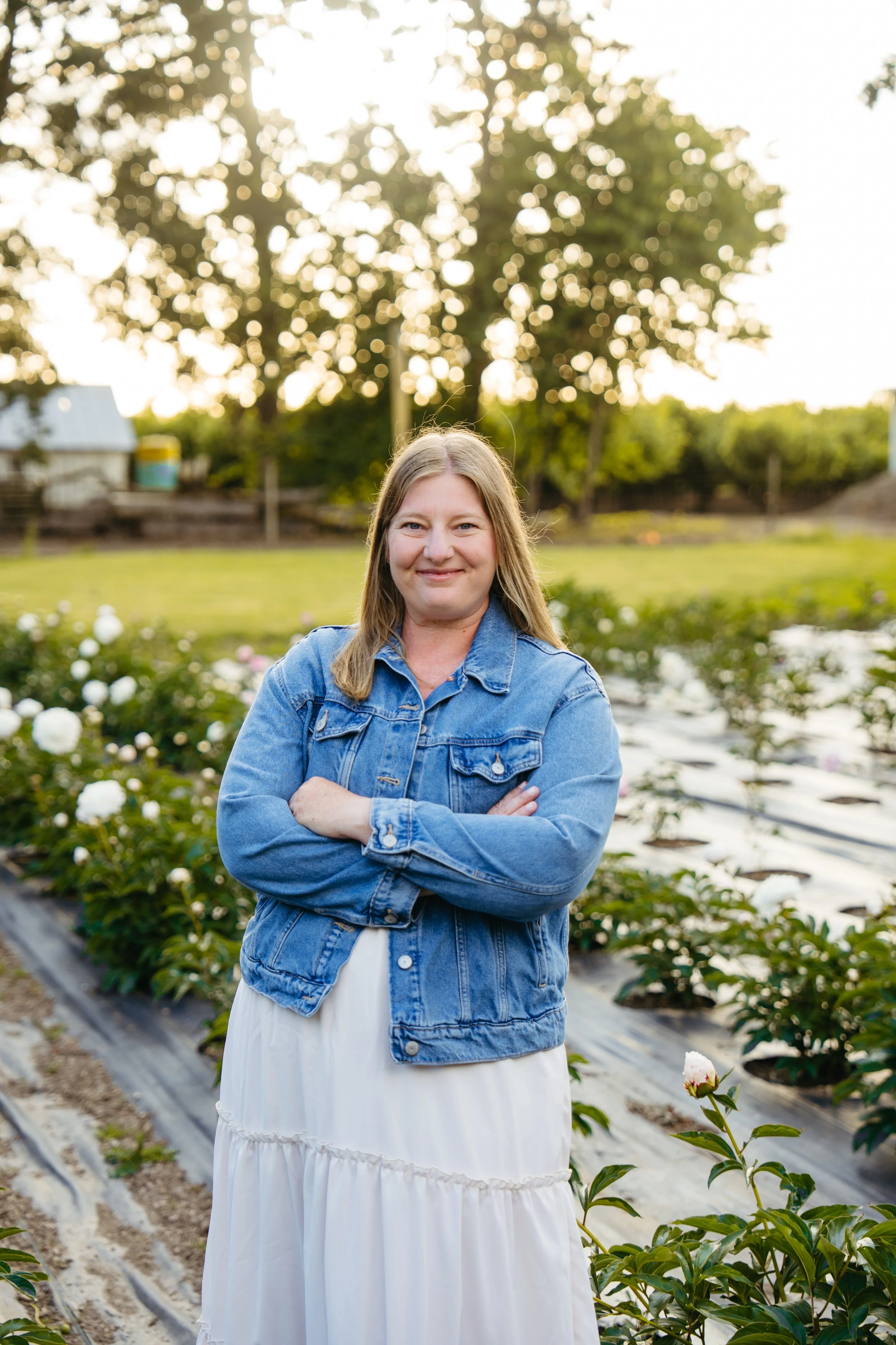 A woman with light brown hair, wearing a denim jacket and a white dress, stands with her arms crossed in a field of blooming white flowers, with a large tree and sunlight in the background.