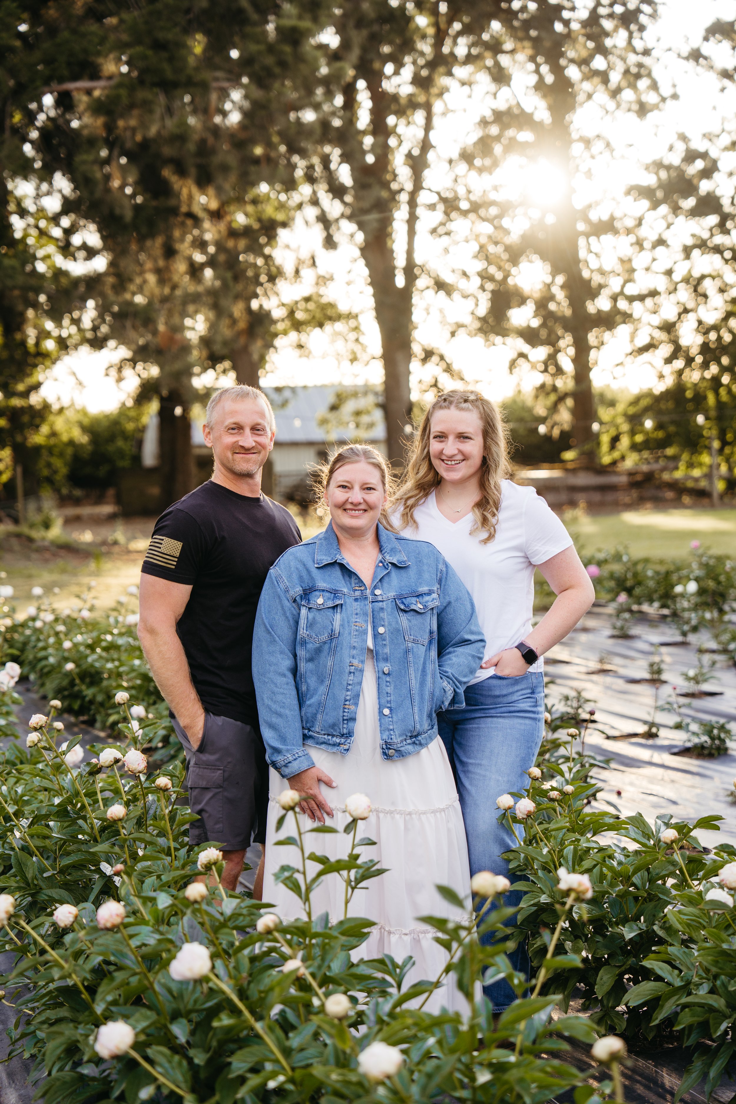 A smiling family of three standing outdoors with trees in the background. The family consists of a young woman in a red dress, a woman in a black sweater, and a man in a green t-shirt and jeans.