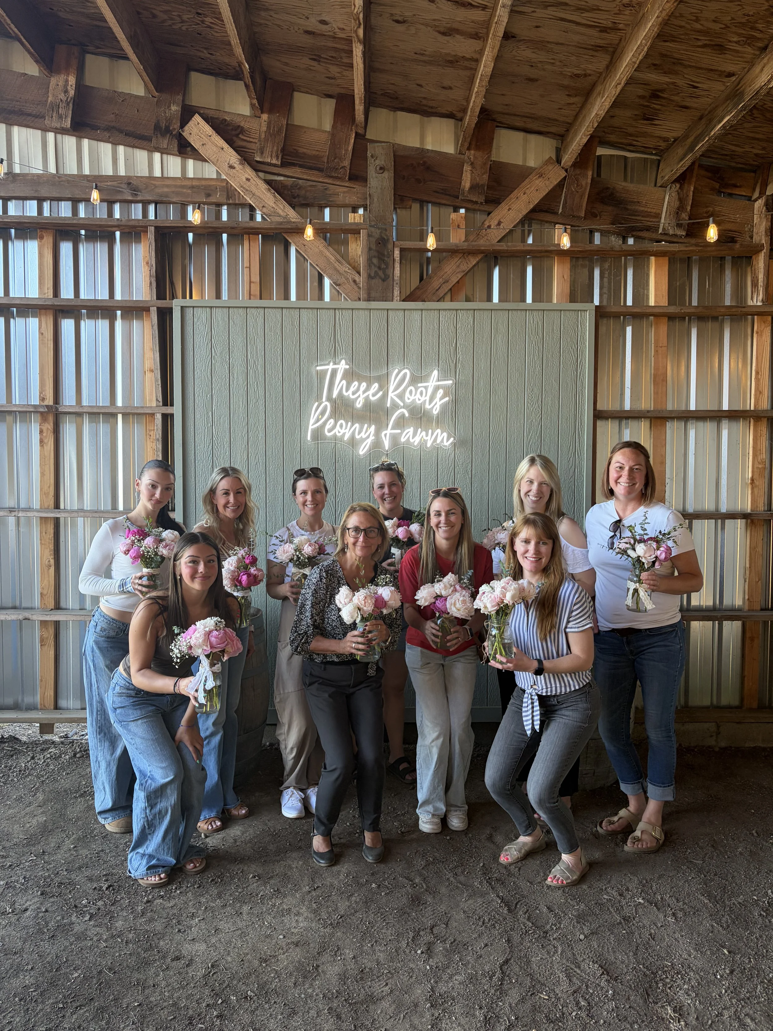 A group of women standing in front of a wooden wall with a sign that says "These Roots Peony Farm." The women are holding bouquets of pink and white peonies and smiling at the camera. The setting appears to be inside a barn or farm structure with a rustic wooden ceiling and hanging string lights.