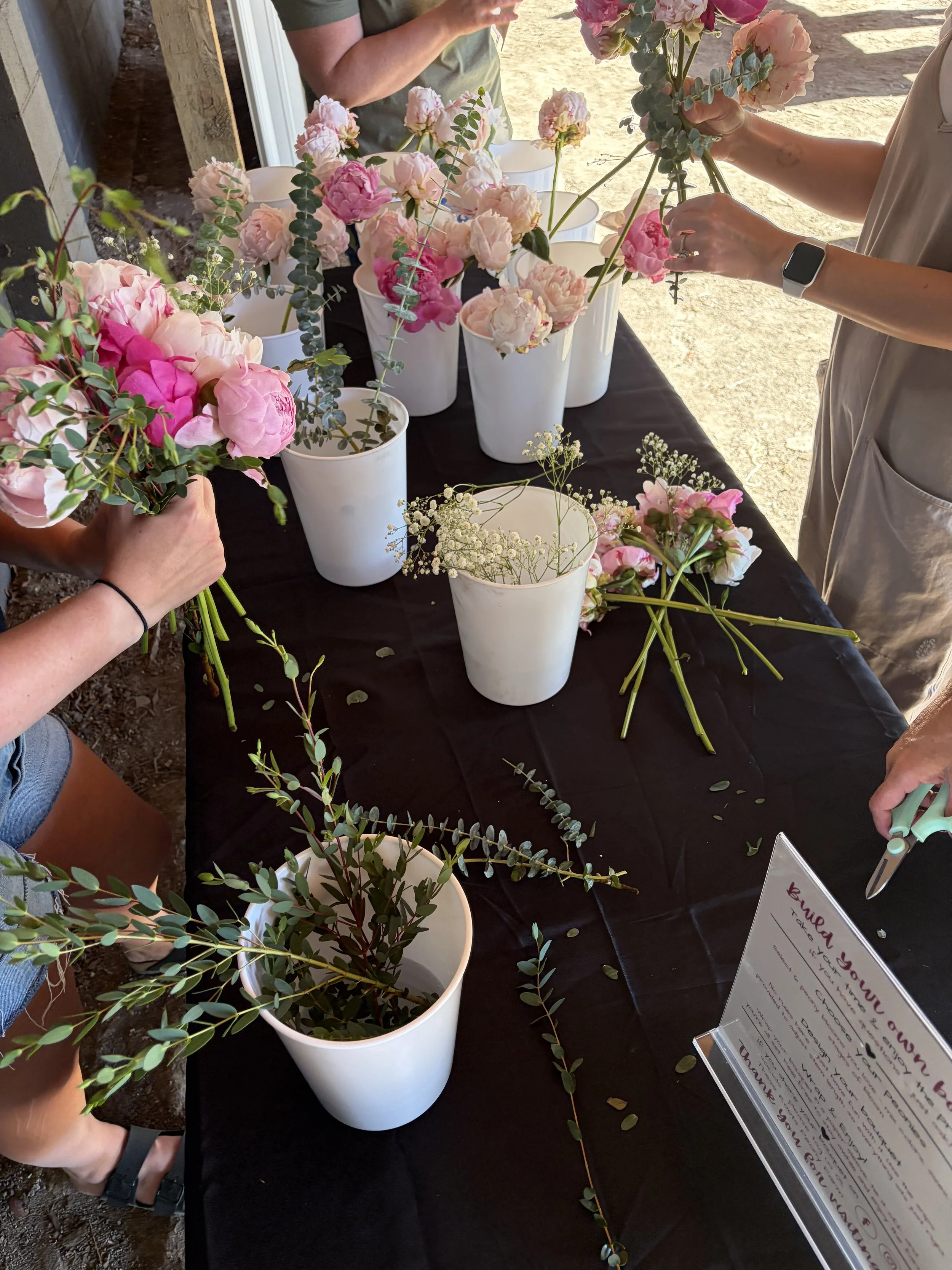 Text overlay on a white wooden background with pink peony flowers on the right side, reading "Girl's Night @ These Roots Peony Farm"