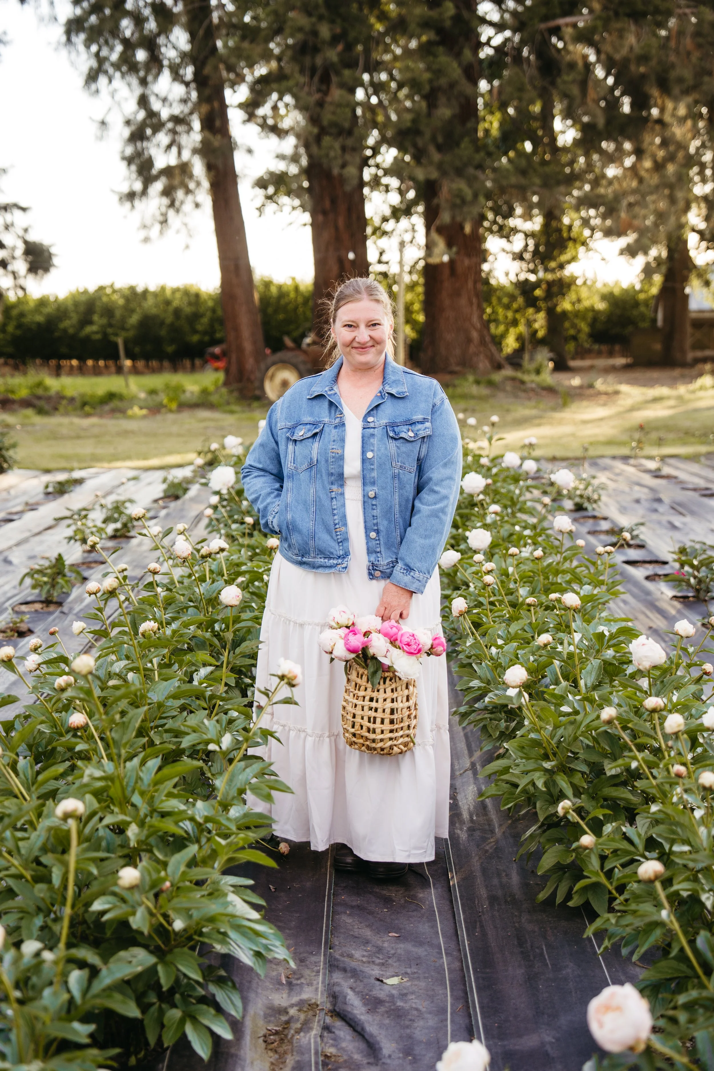 A woman standing in a flower farm holding a basket of pink and white flowers, with greenery and large trees in the background, during daylight.