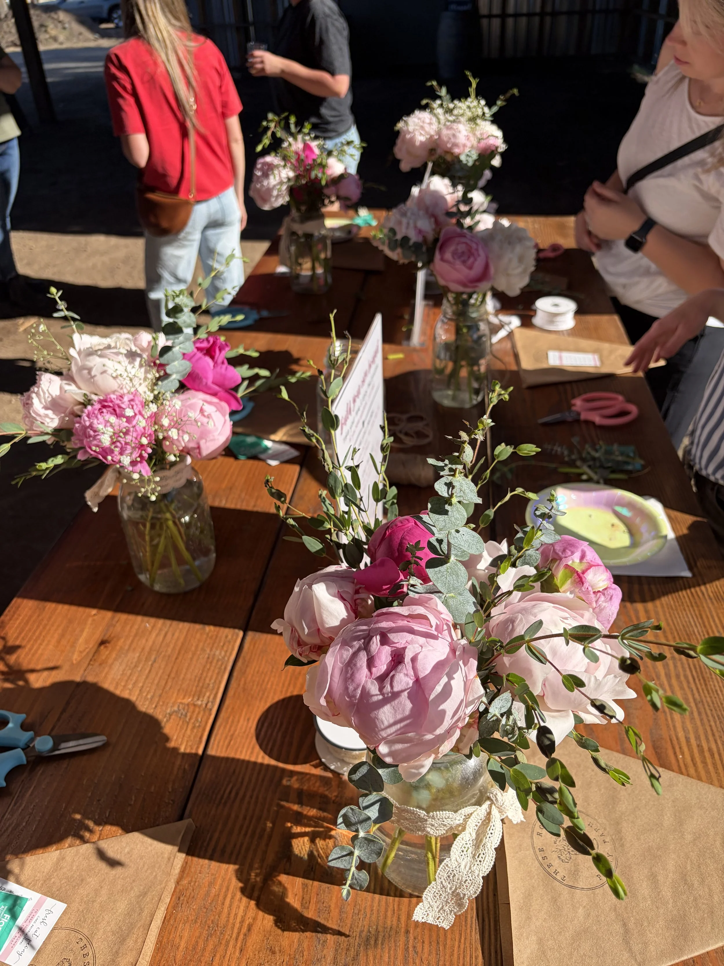 Various pink and white flowers in glass jars on a wooden table with people in the background.