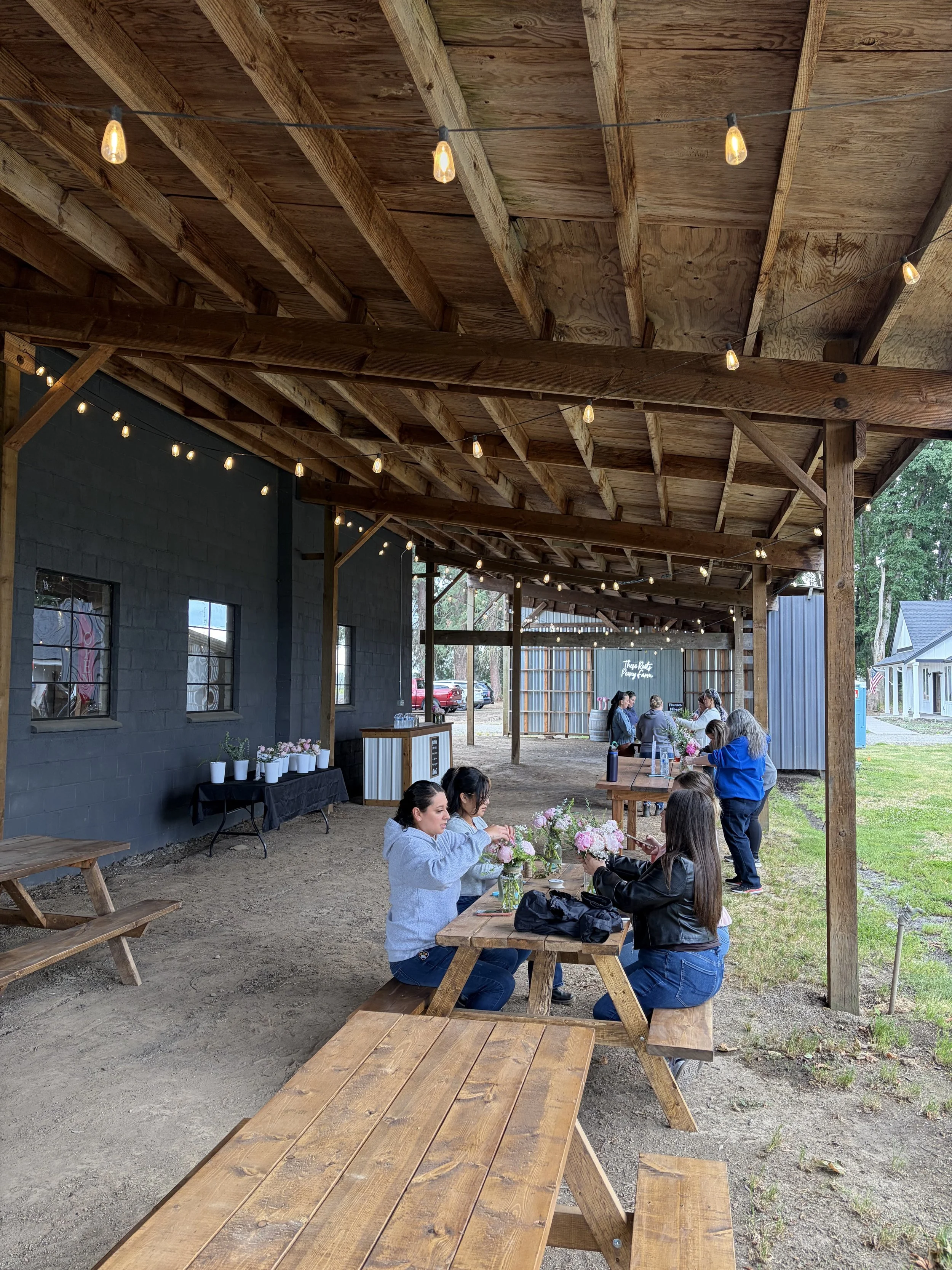 People seated at picnic tables under a wooden pavilion decorated with string lights, with flowers on the tables and a building in the background.