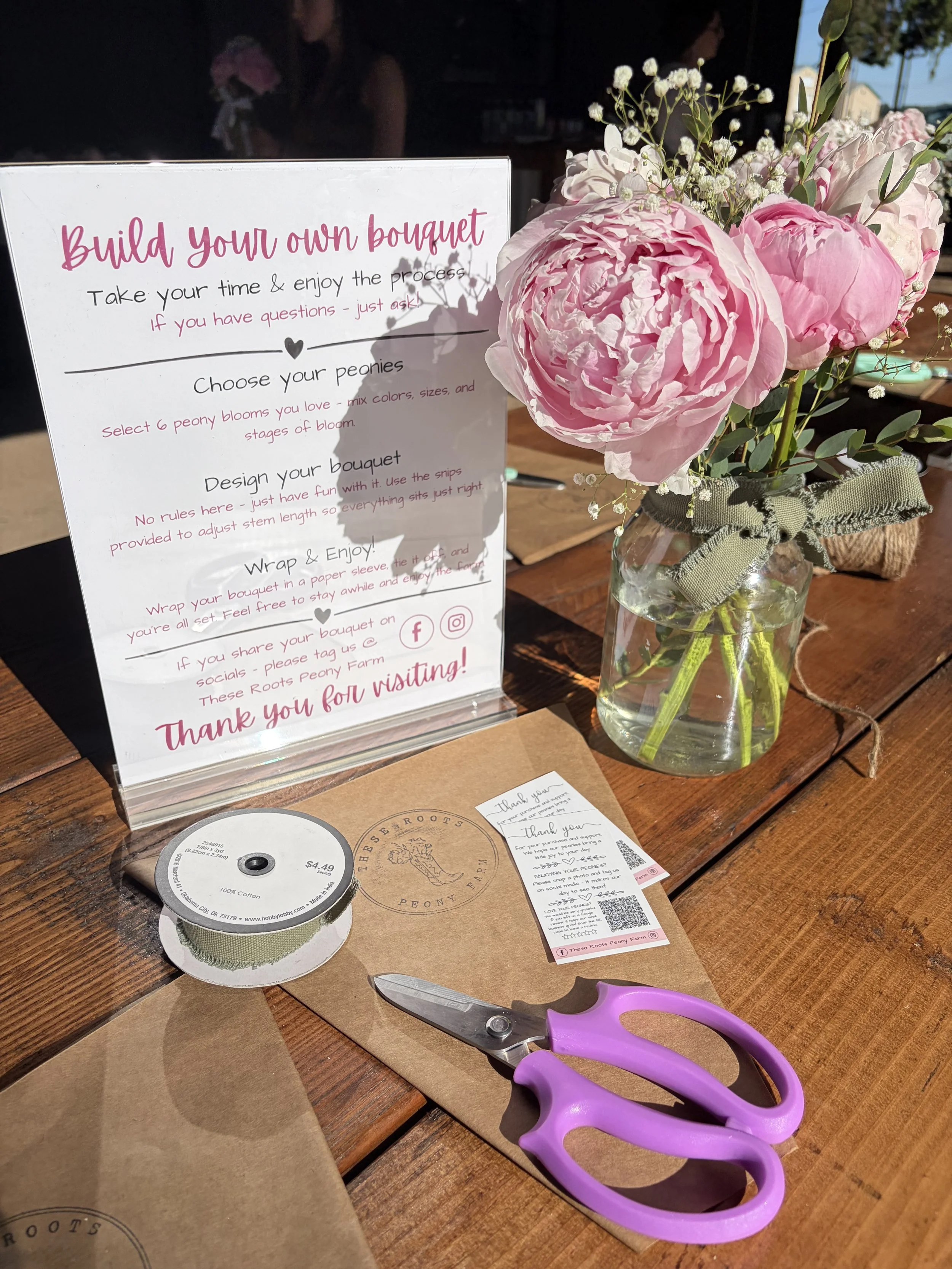 A table with craft supplies for flower bouquet making, including pink scissors, brown paper, a roll of ribbon, a glass jar with pink and white flowers, and printed instructions.