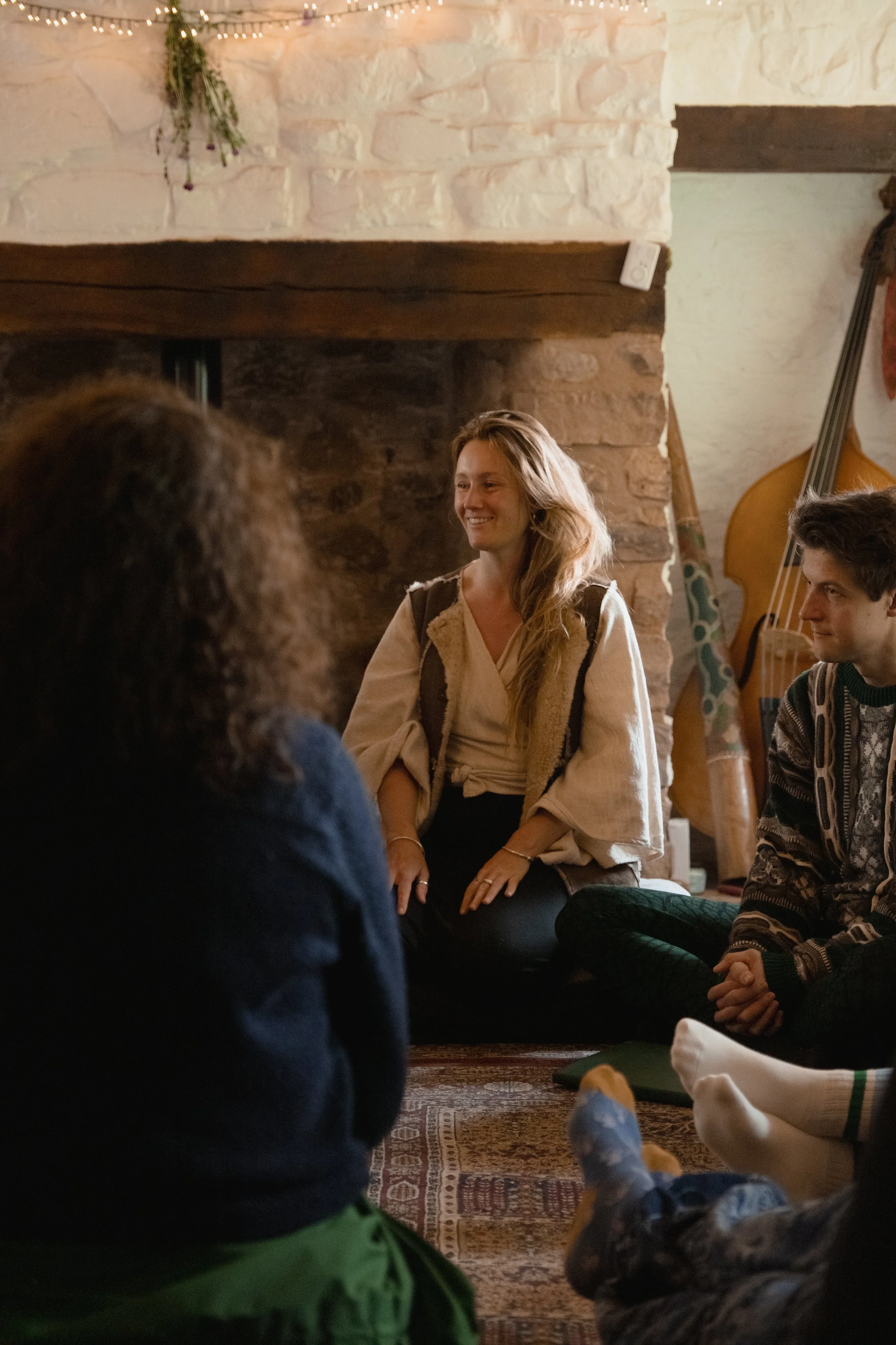 A woman with long blonde hair, wearing a beige top and vest, smiling while sitting on a rug. Around her, people are seated on the floor. The room features a stone fireplace with a wooden beam and a double bass leaning against the wall.