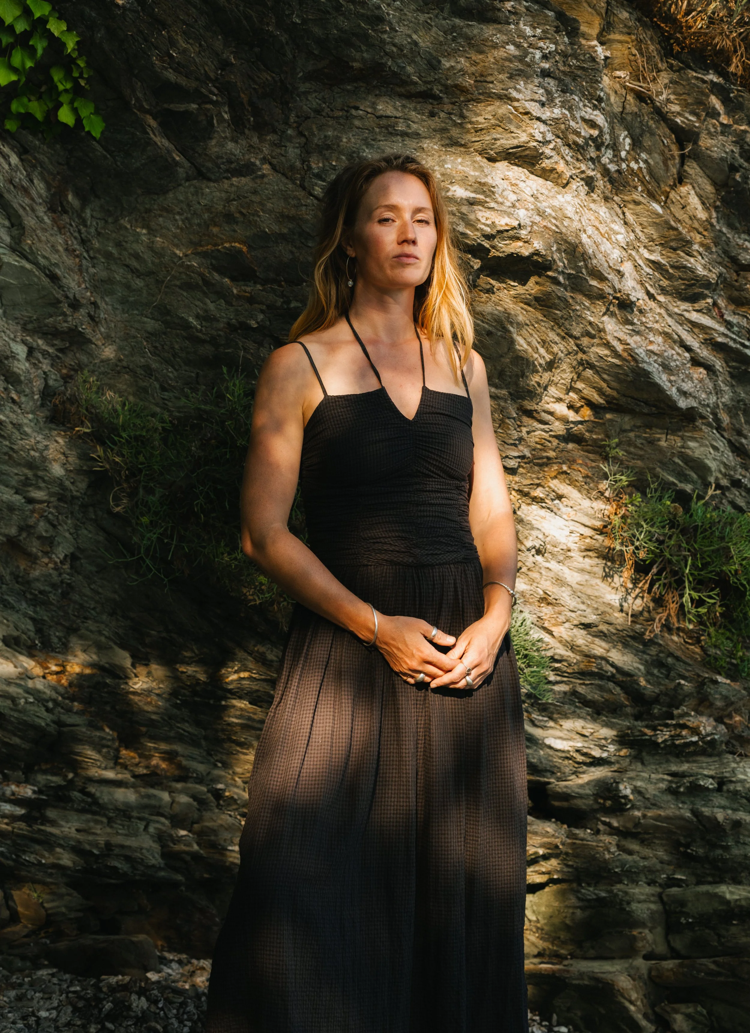 A woman with long, wavy, light brown hair wearing a black spaghetti strap top and brown patterned skirt standing outdoors against a rocky background with some green plants.