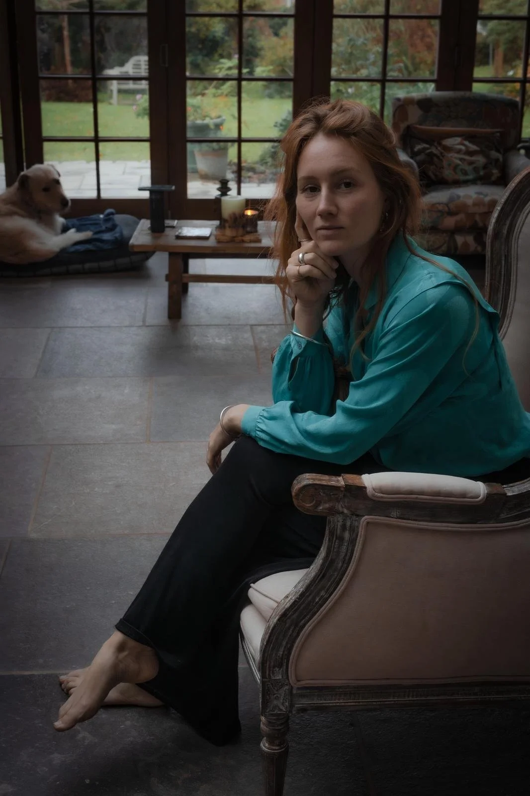 A woman with red hair in a blue satin blouse sitting on a vintage armchair visible in a sunroom or enclosed porch, with a dog lying on a cushion on a bench nearby. There is a wooden table with lamps and candles in the background, and large windows overlooking a garden with greenery and a patio.