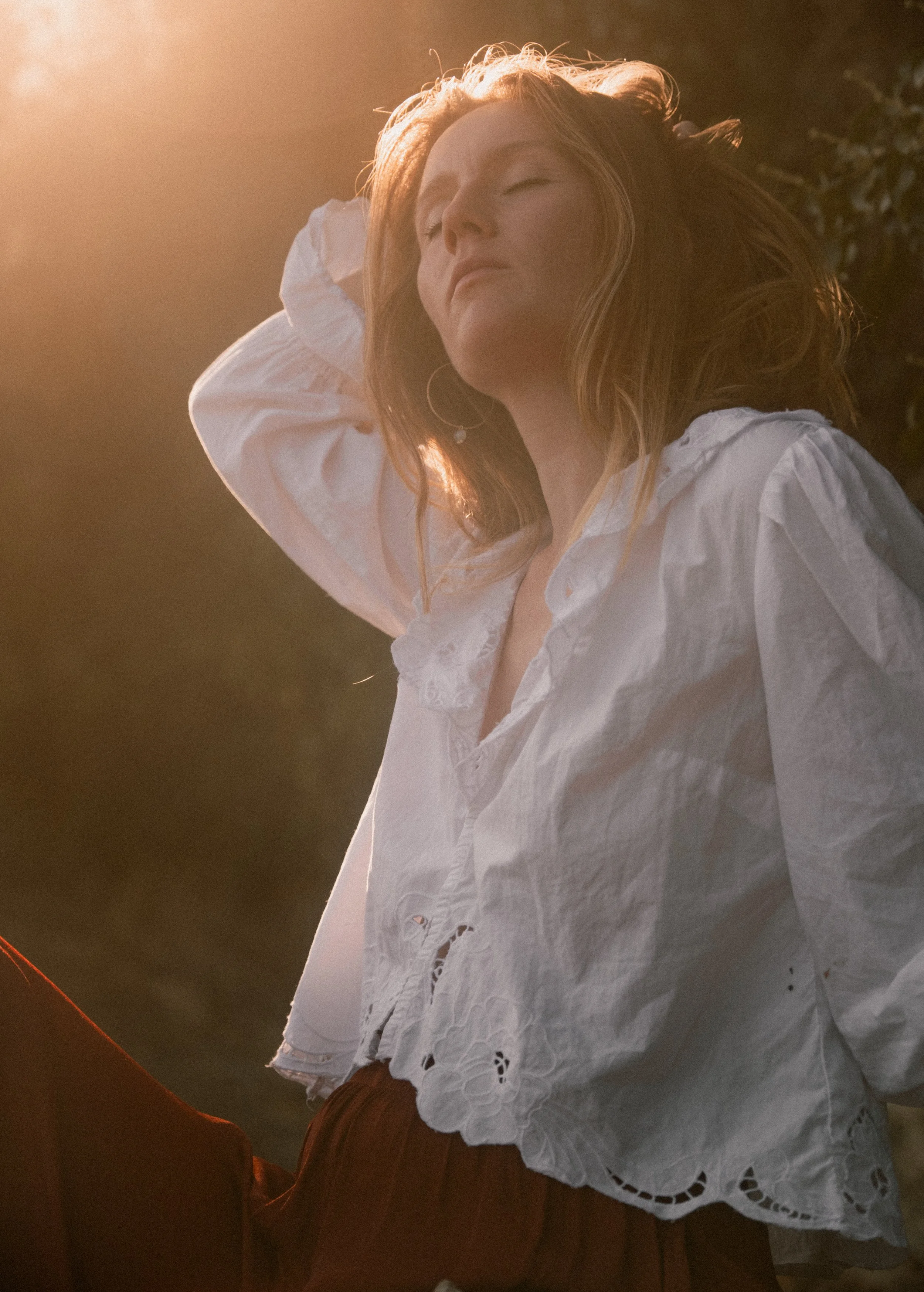 A woman with red hair and light skin lying outdoors with her eyes closed, wearing a white blouse with lace details and a white hat, in warm sunlight.