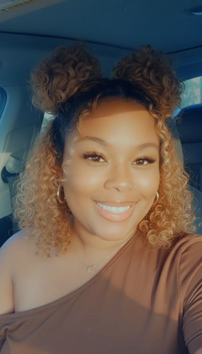 A woman with curly, two-toned hair in a bun on top of her head, smiling at camera, inside a vehicle.