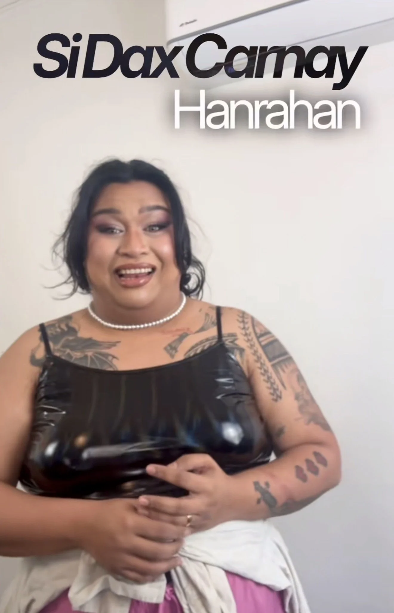 A person with dark hair, tattoos on arms, wearing a black leather top, holding a camera, standing in front of a white wall with text that reads 'siDax Canay Hanrahan'.