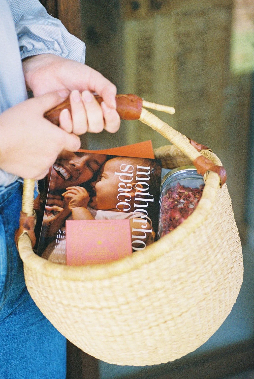 Person holding a woven basket containing a book, a pink card, and a jar of dried rose petals.