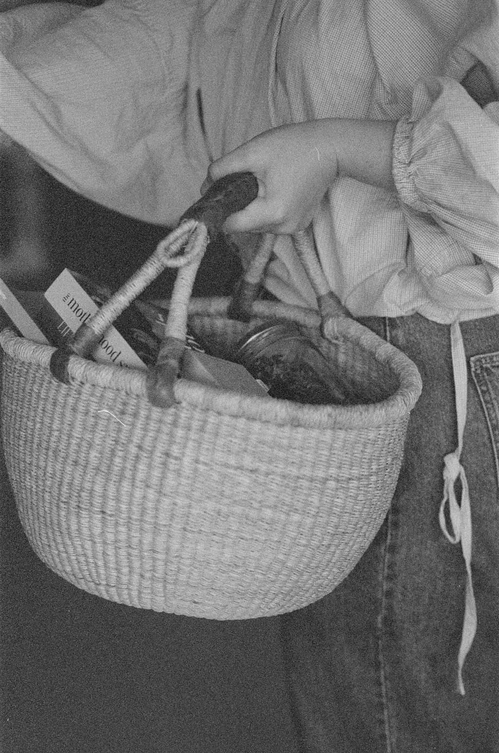 A person wearing a long-sleeved shirt is reaching into a woven basket filled with objects, including a bottle and a book, while sitting on a textured surface.