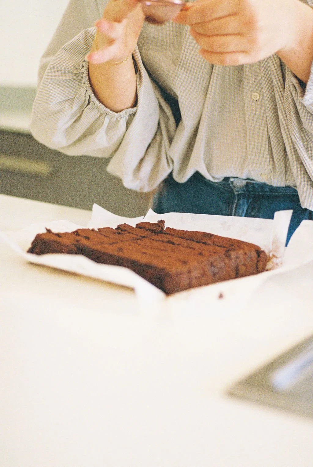 Person sprinkling powdered sugar on a large chocolate bar on a white paper-lined tray.