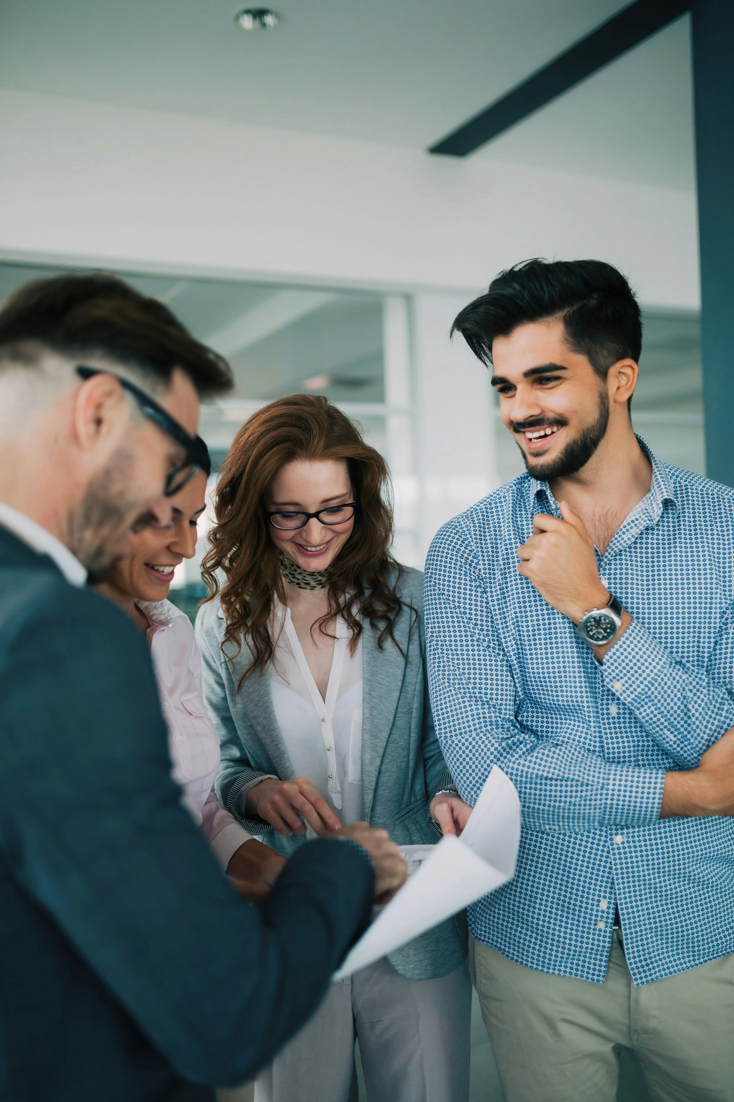 Group of four diverse professionals gathered around, smiling, and looking at documents in a modern office.