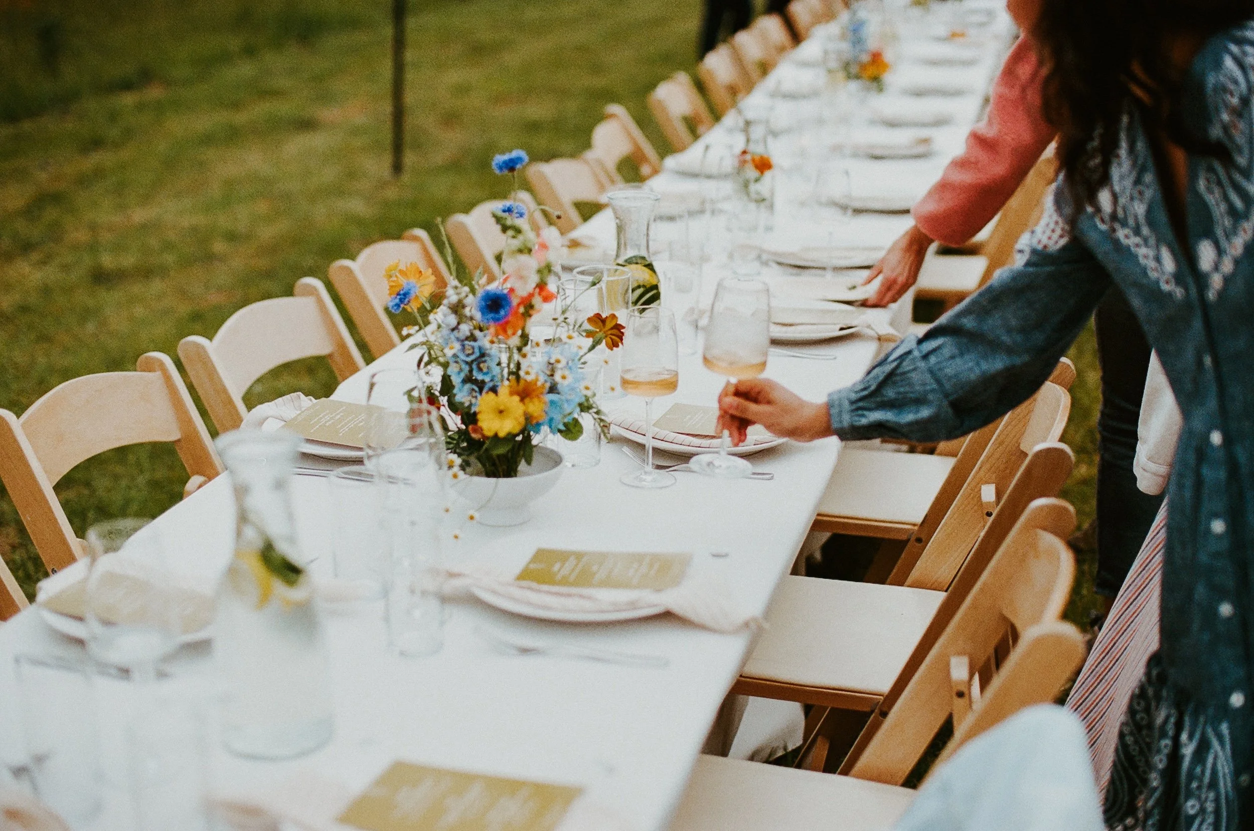Long outdoor table set for a social event, decorated with colorful flower arrangements, white tablecloth, plates, wine glasses, and utensils. Several people are seen arranging the table, which is situated on a grassy outdoor area.