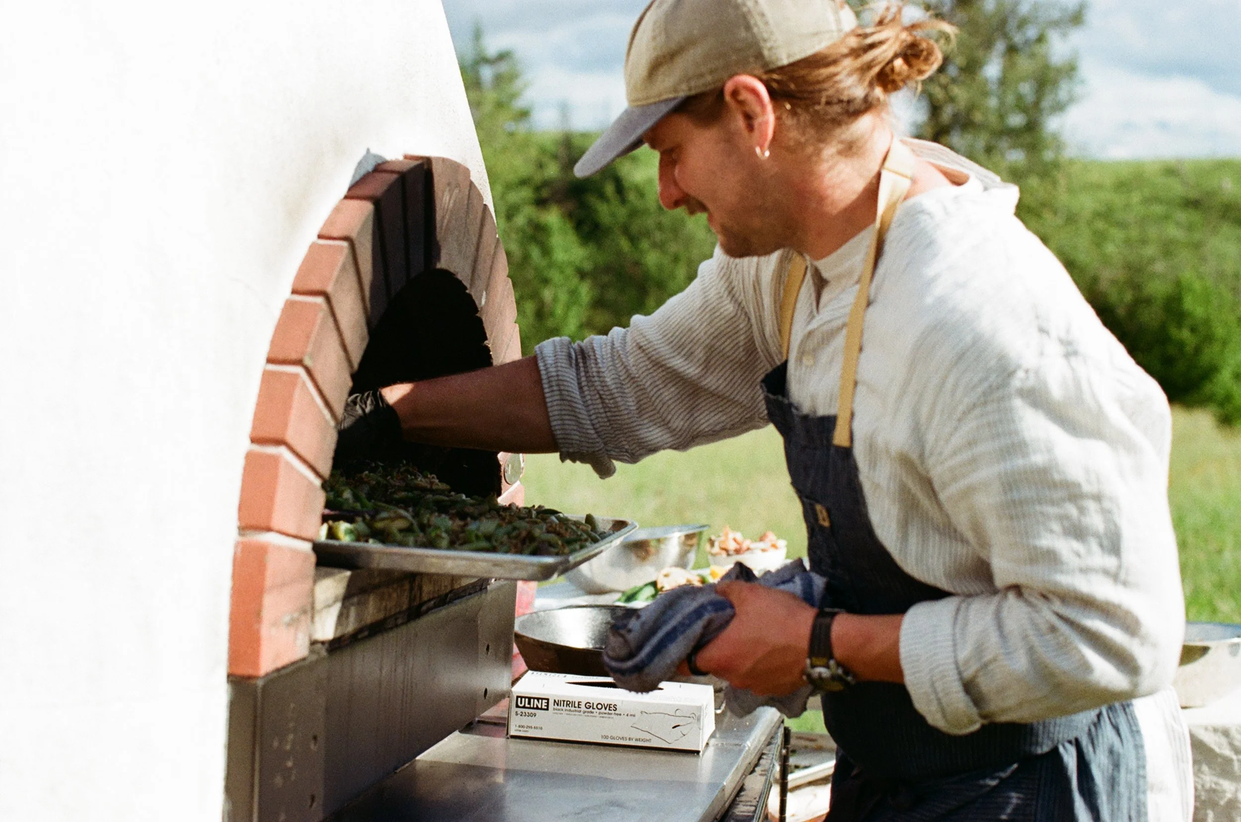 A man wearing a beige cap and black gloves is preparing food outdoors. He is leaning over a wooden table with a brick oven in the background, surrounded by green trees and a partly cloudy sky.