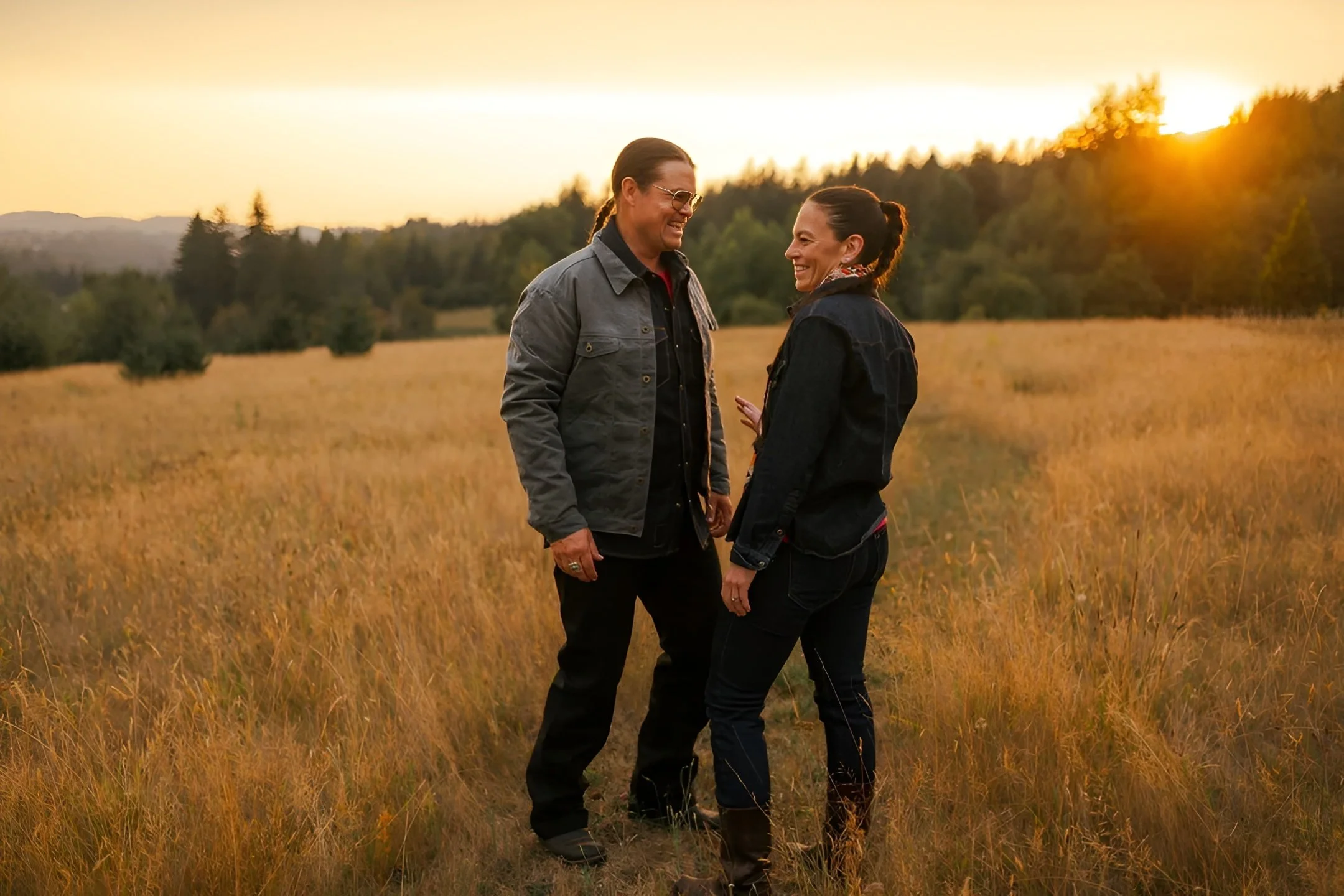 A man and woman standing in a grassy field during sunset, smiling at each other.