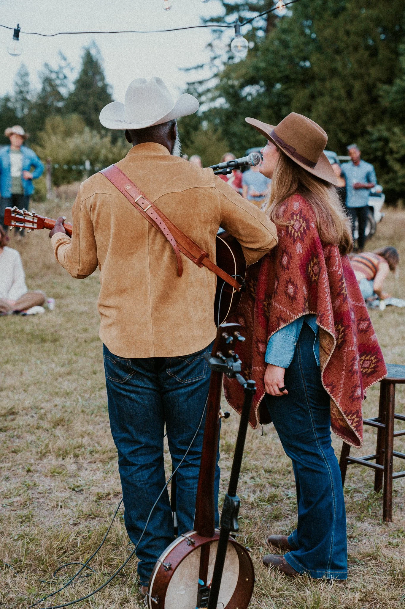 A man playing guitar and a woman singing into a microphone outdoors, wearing cowboy hats, with string lights overhead and a group of people in the background.