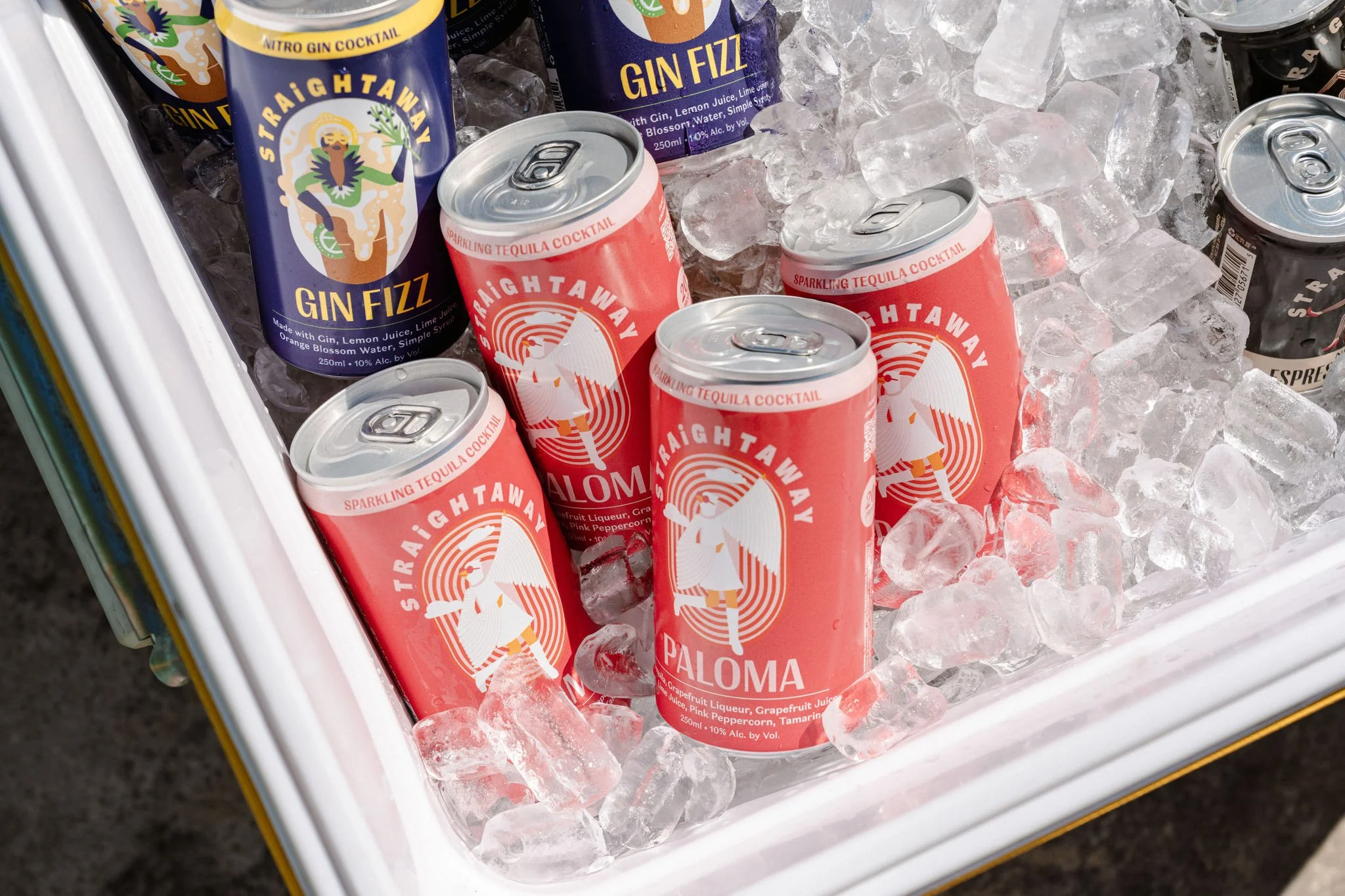 A cooler filled with ice containing cans of drinks, including pink cans labeled 'Paloma' and blue cans labeled 'Gin Fizz' with colorful designs and text.