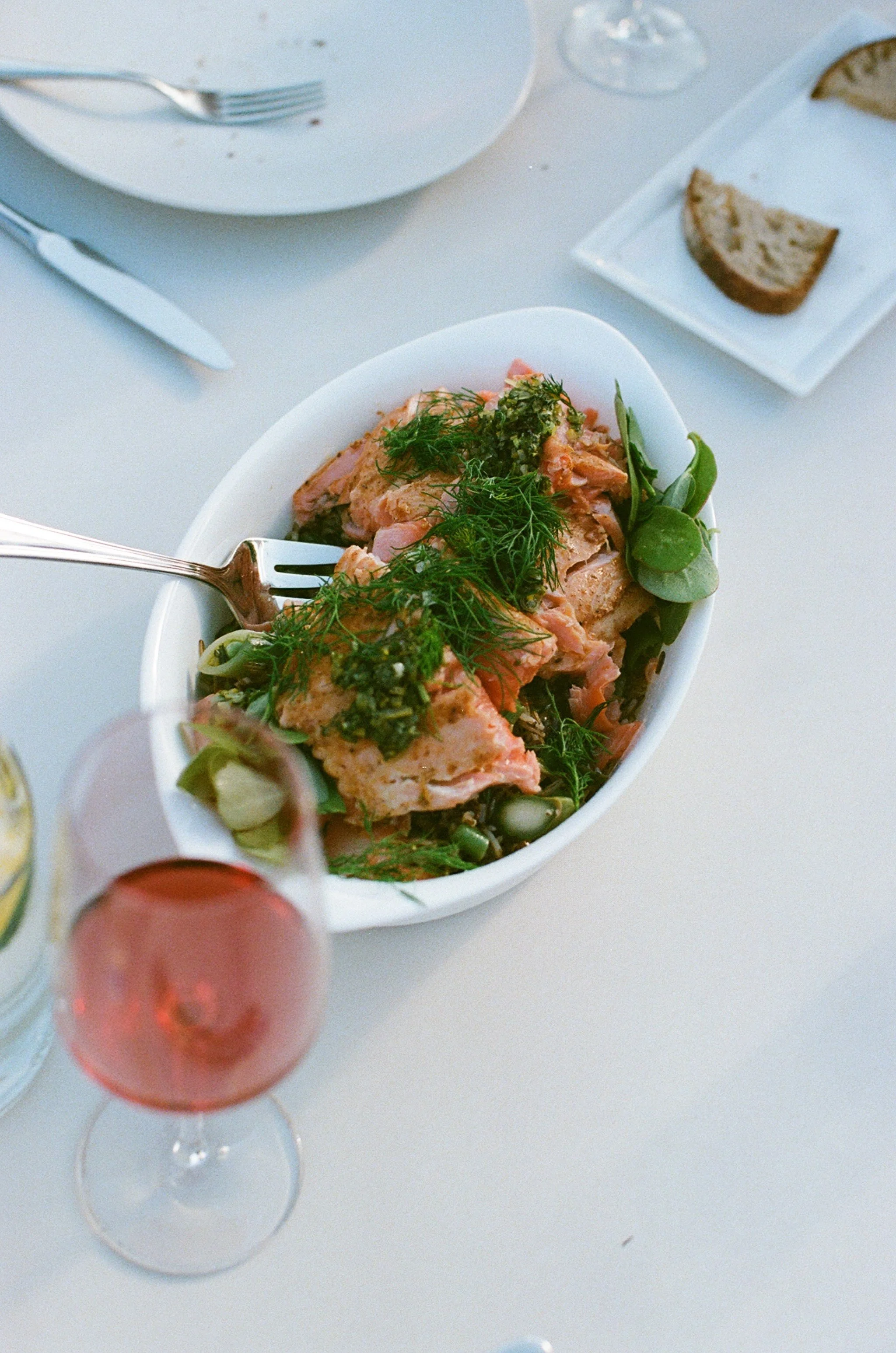 A white dish containing a salad with herbs, served with slices of bread on a white plate. In the foreground, a glass of pinkish red wine is partially visible. The setting appears to be a meal on a white table, with a fork, knife, and empty plate in the background.