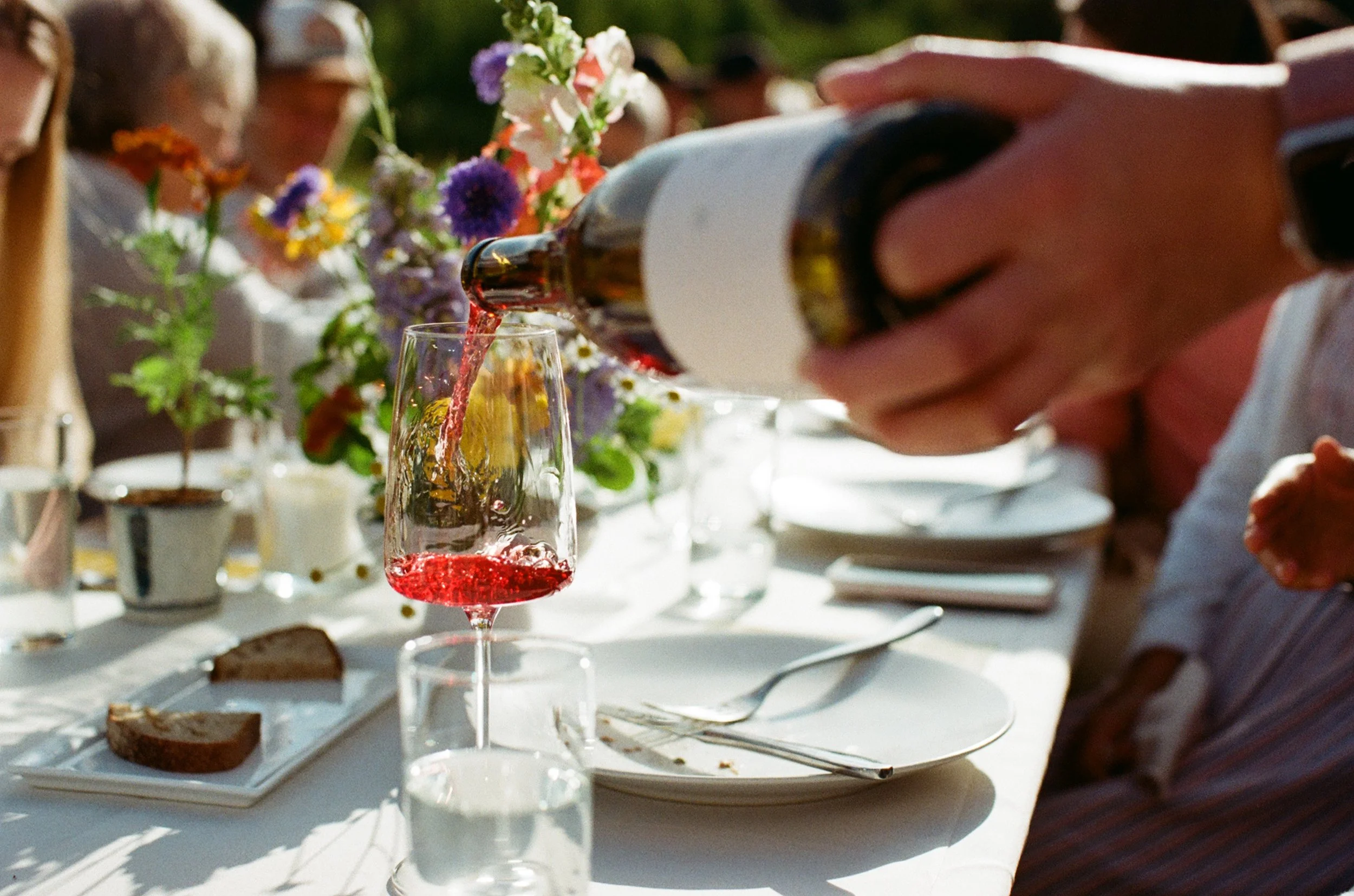A person is pouring red wine into a glass on a dinner table decorated with flowers and food. There are other glasses and plates on the table with a blurred outdoor setting in the background.