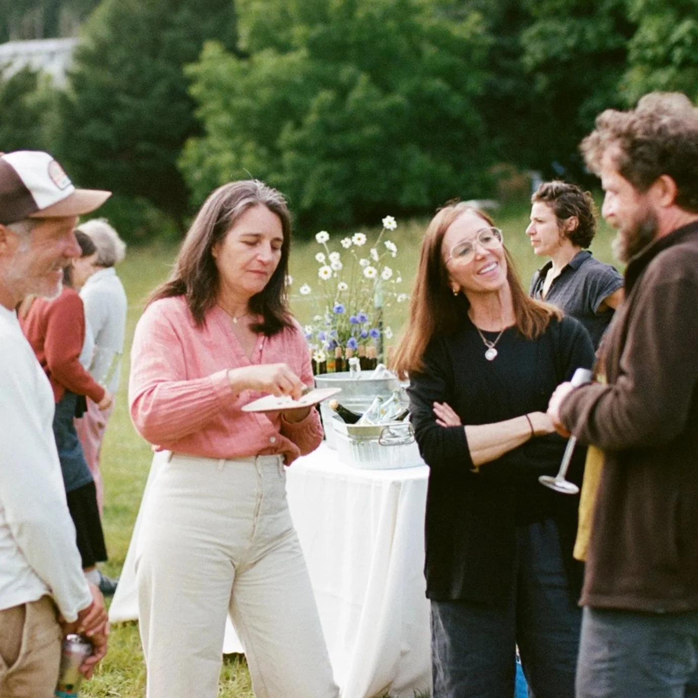 A group of people gathering outdoors at a social event with a table of flowers and drinks, engaging in conversation.