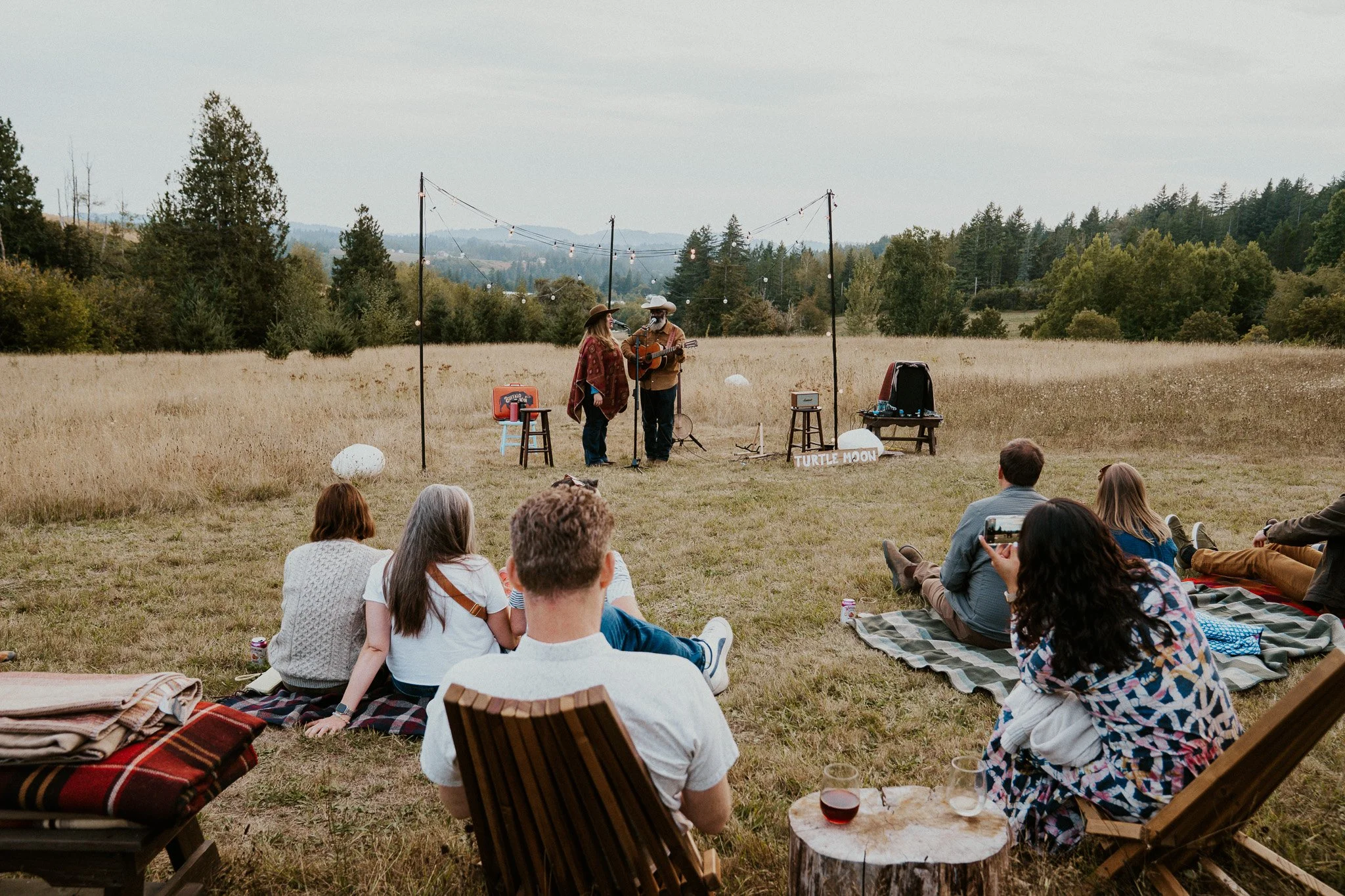 People sitting on blankets and chairs in an open field, attending an outdoor acoustic music performance under string lights, with musicians wearing hats in the background and trees in the distance.
