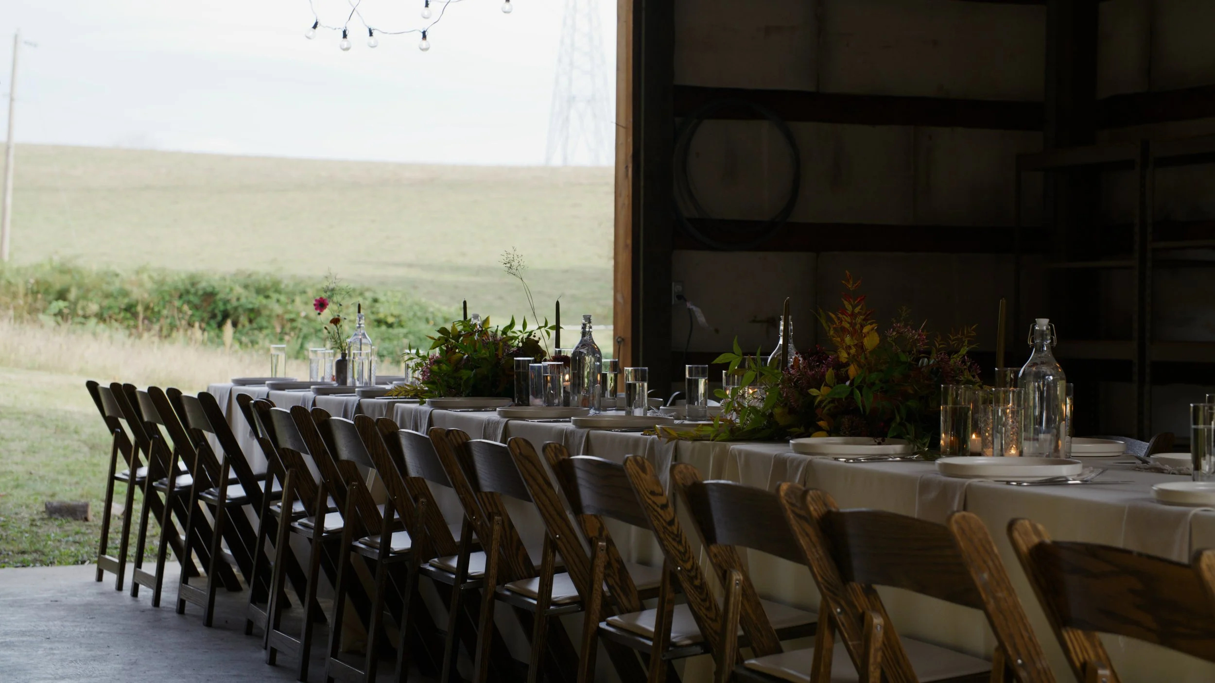 Long banquet table set with white tablecloths, glass bottles, glasses, plates, and floral centerpieces inside a rustic venue. Outside, a grassy field and distant trees are visible through large open doors.