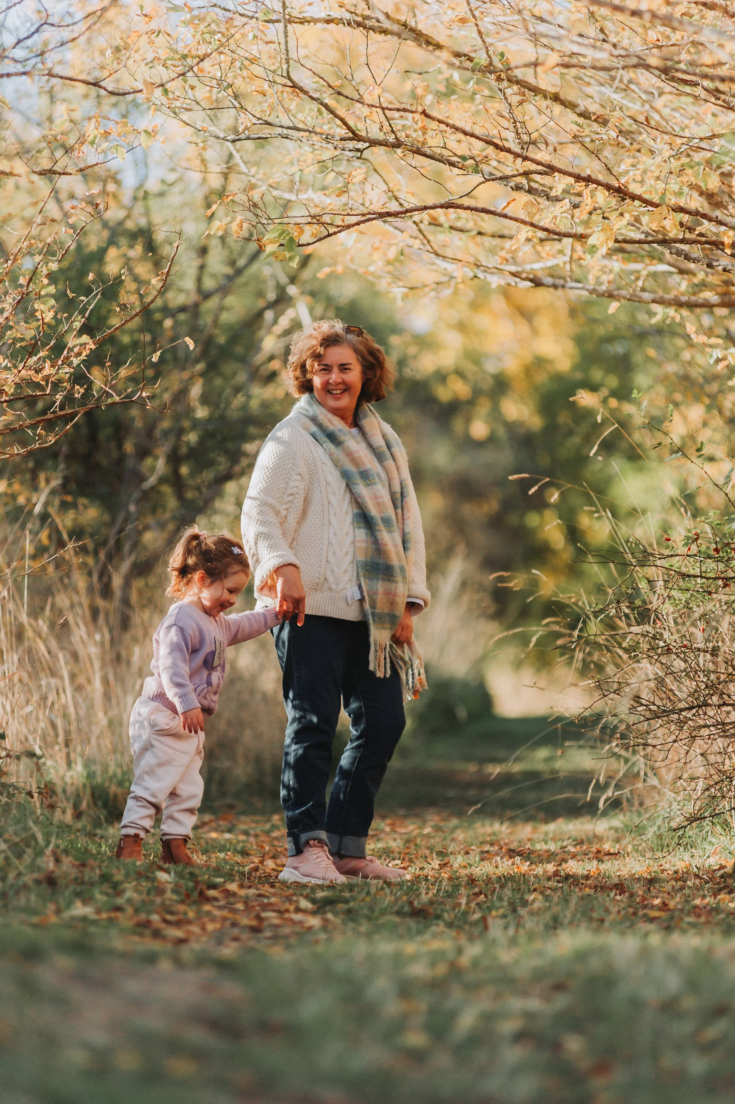 Woman and young girl holding hands and walking on a path surrounded by autumn foliage.