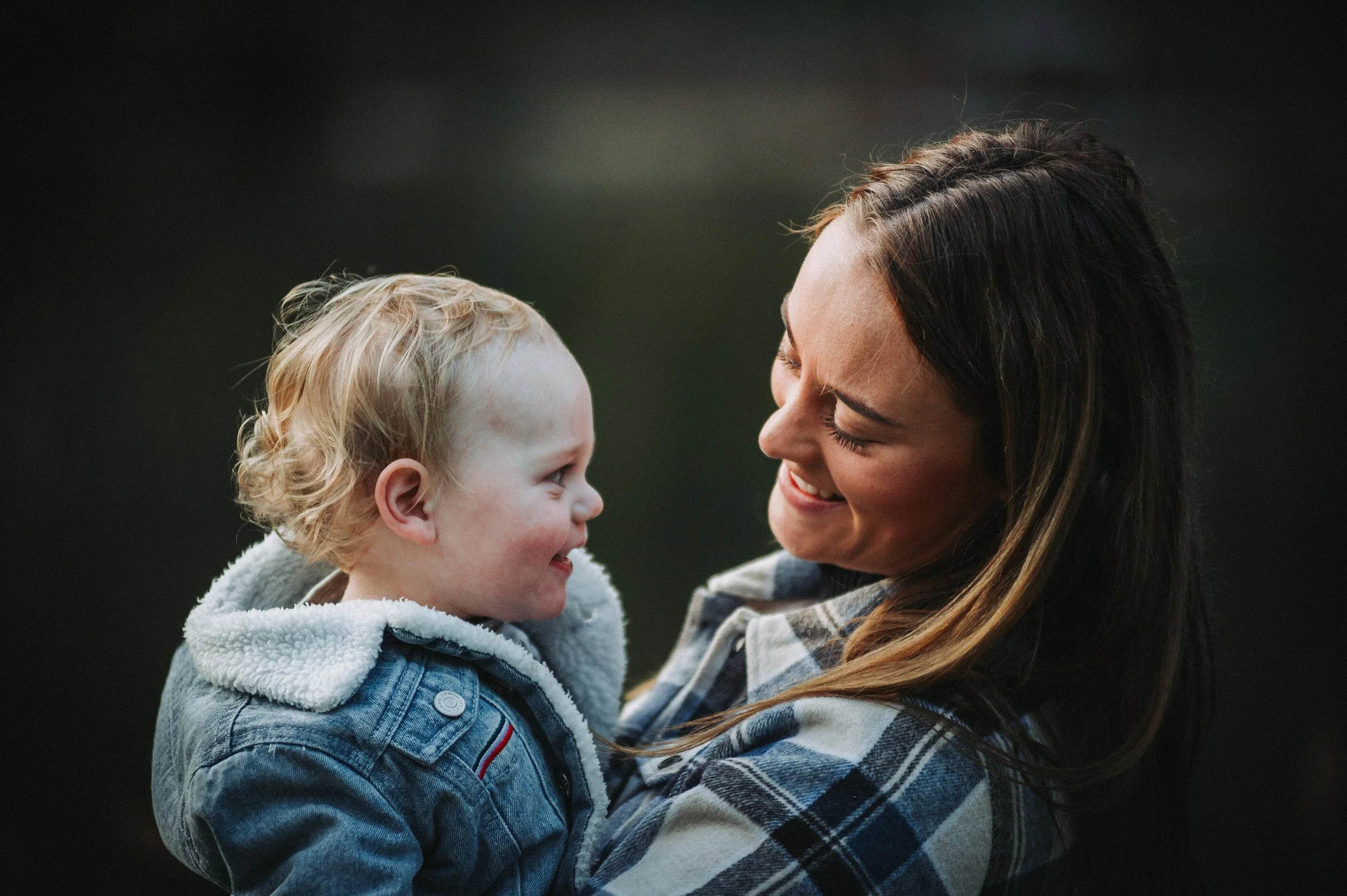 A smiling woman holding a happy child wearing a denim jacket, both sharing a joyful moment together.