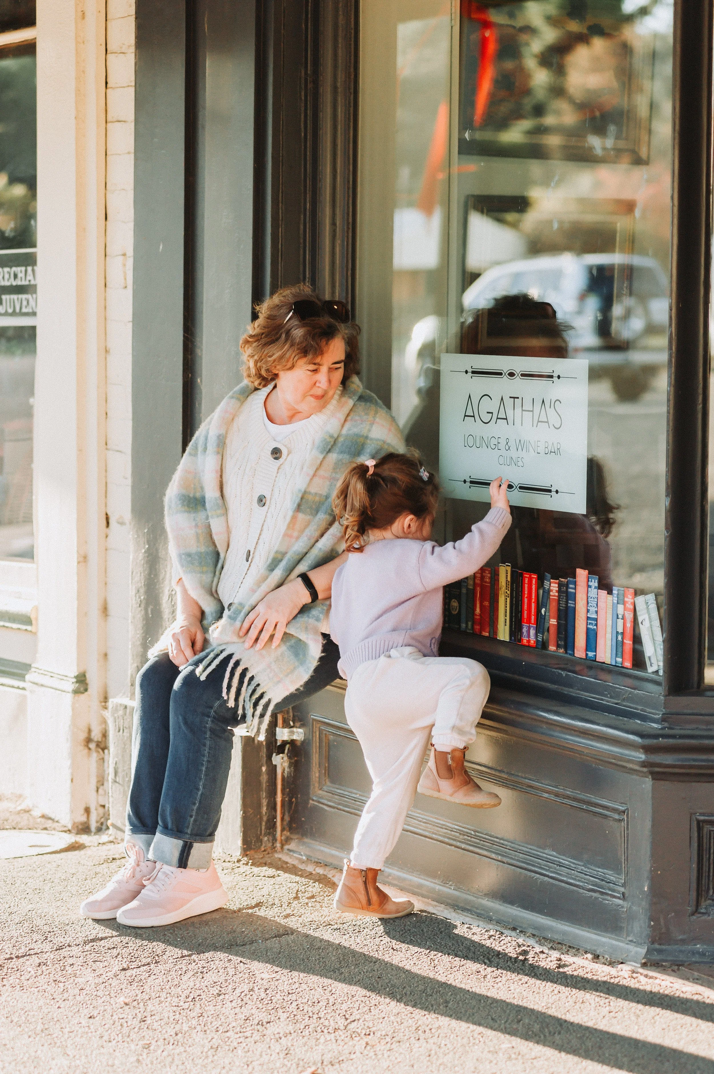 Older woman sitting on ledge next to child browsing books, outside Agatha's Lounge & Wine Bar.